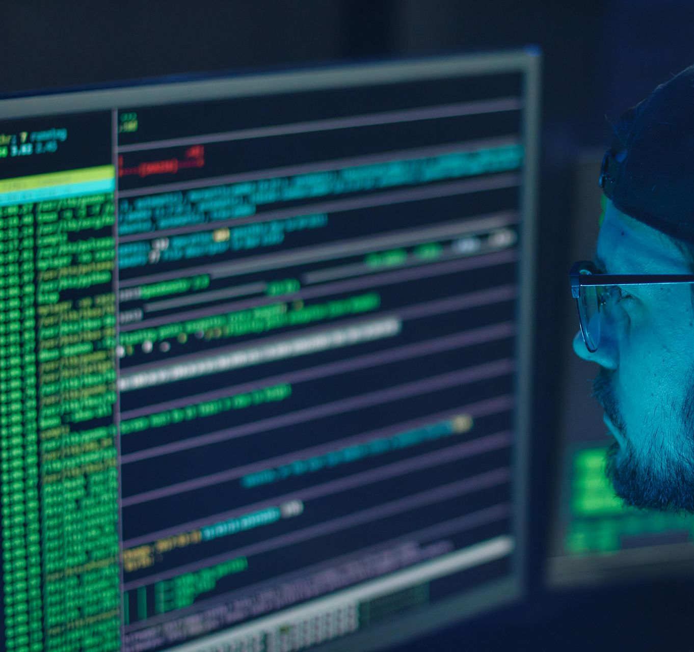 Man in cap and glasses looks at computer screens displaying code, lit by blue light.