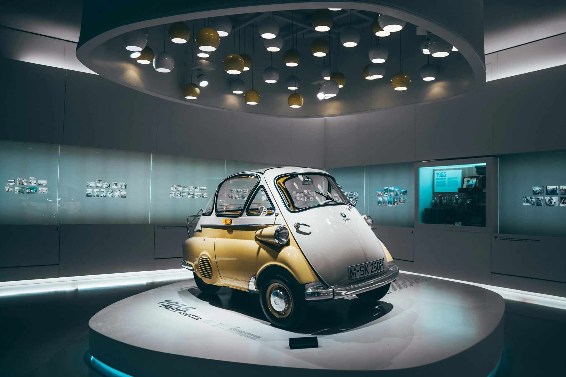 Yellow and white BMW Isetta bubble car on a circular platform in a museum setting, under decorative lights.