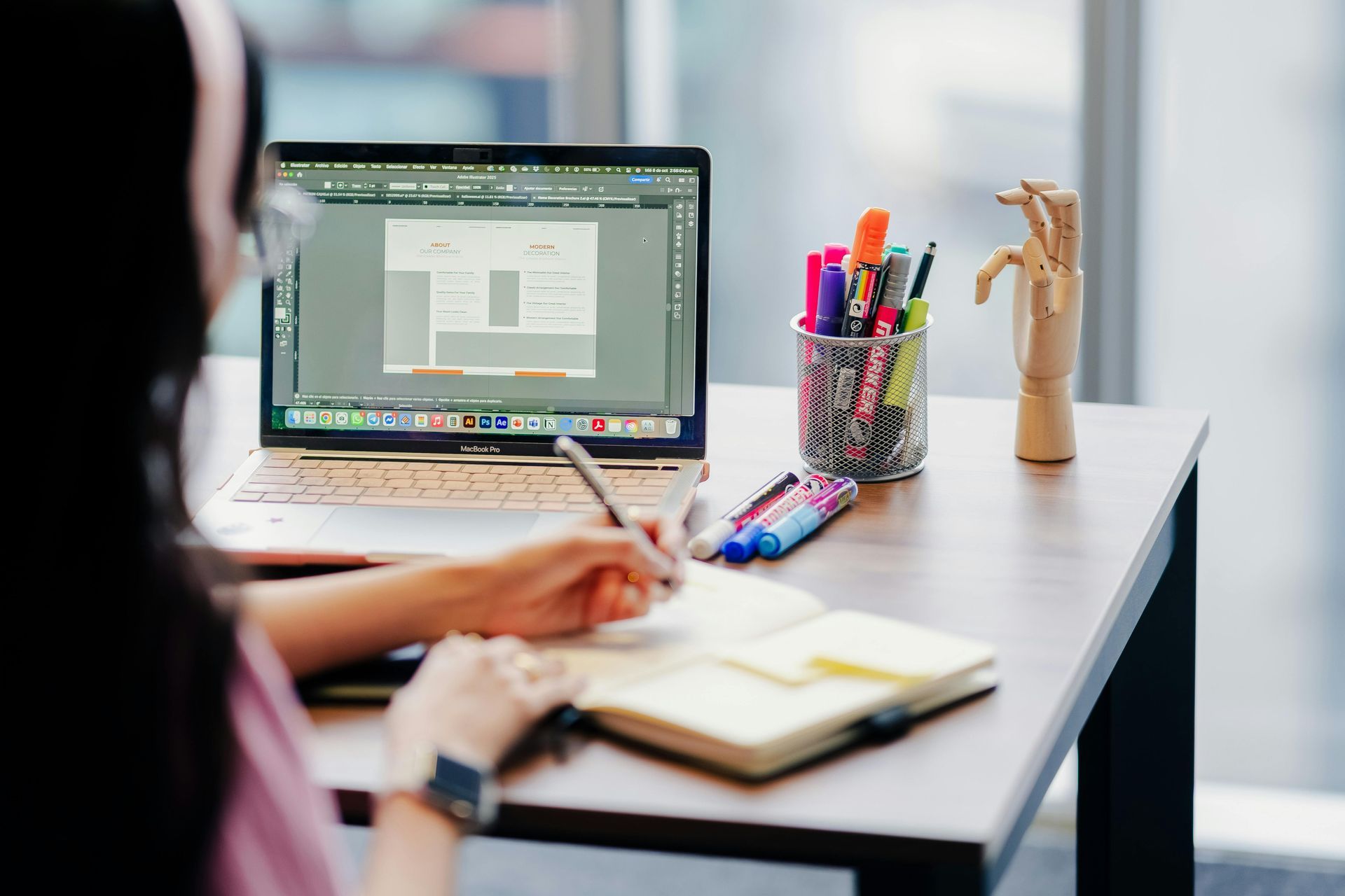 Person working at a desk, looking at a laptop screen. They are writing in a notebook next to a pencil holder.