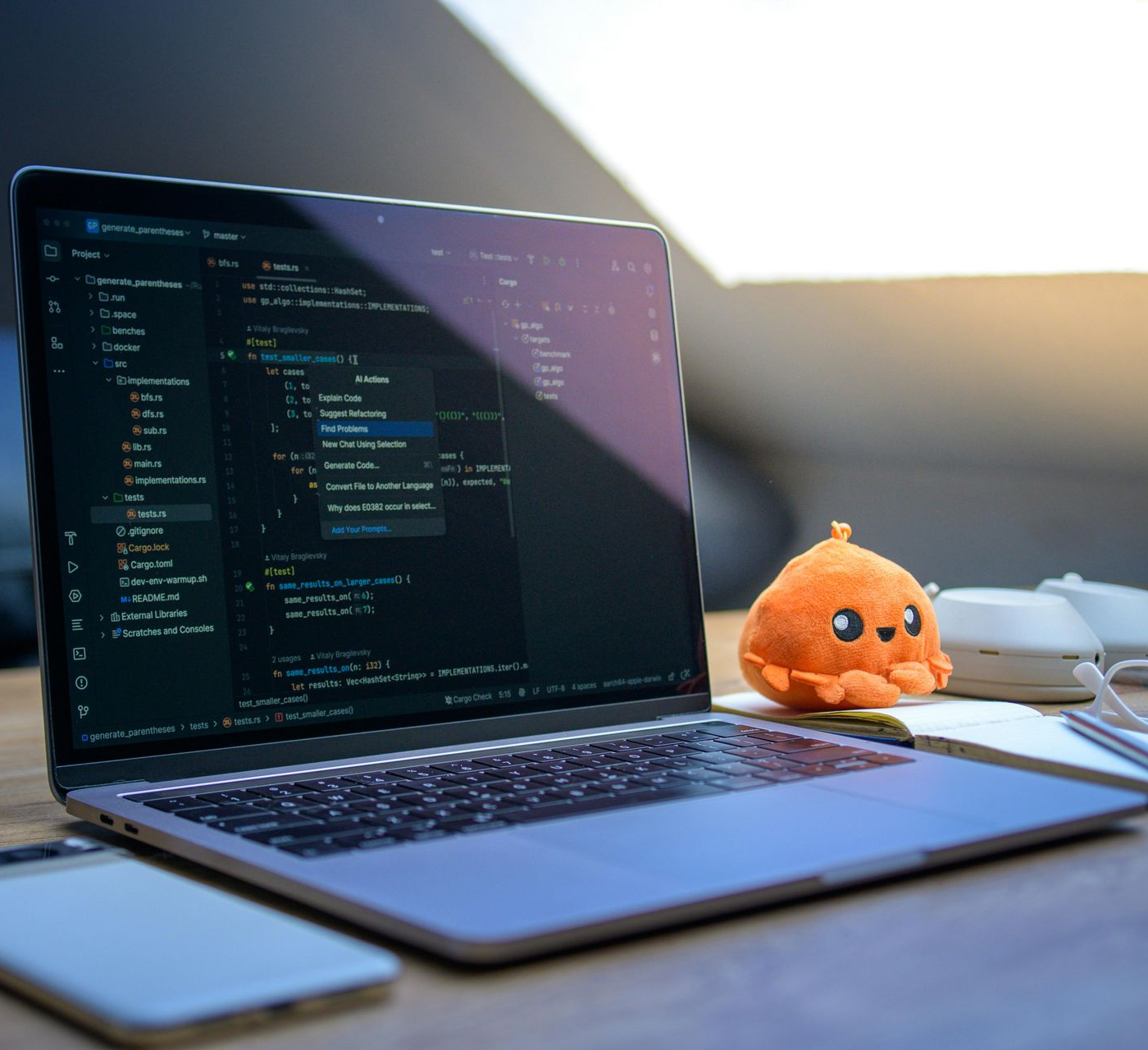 Laptop displaying code, with a stuffed orange octopus, notebook, glasses, and phone on a desk near a window.
