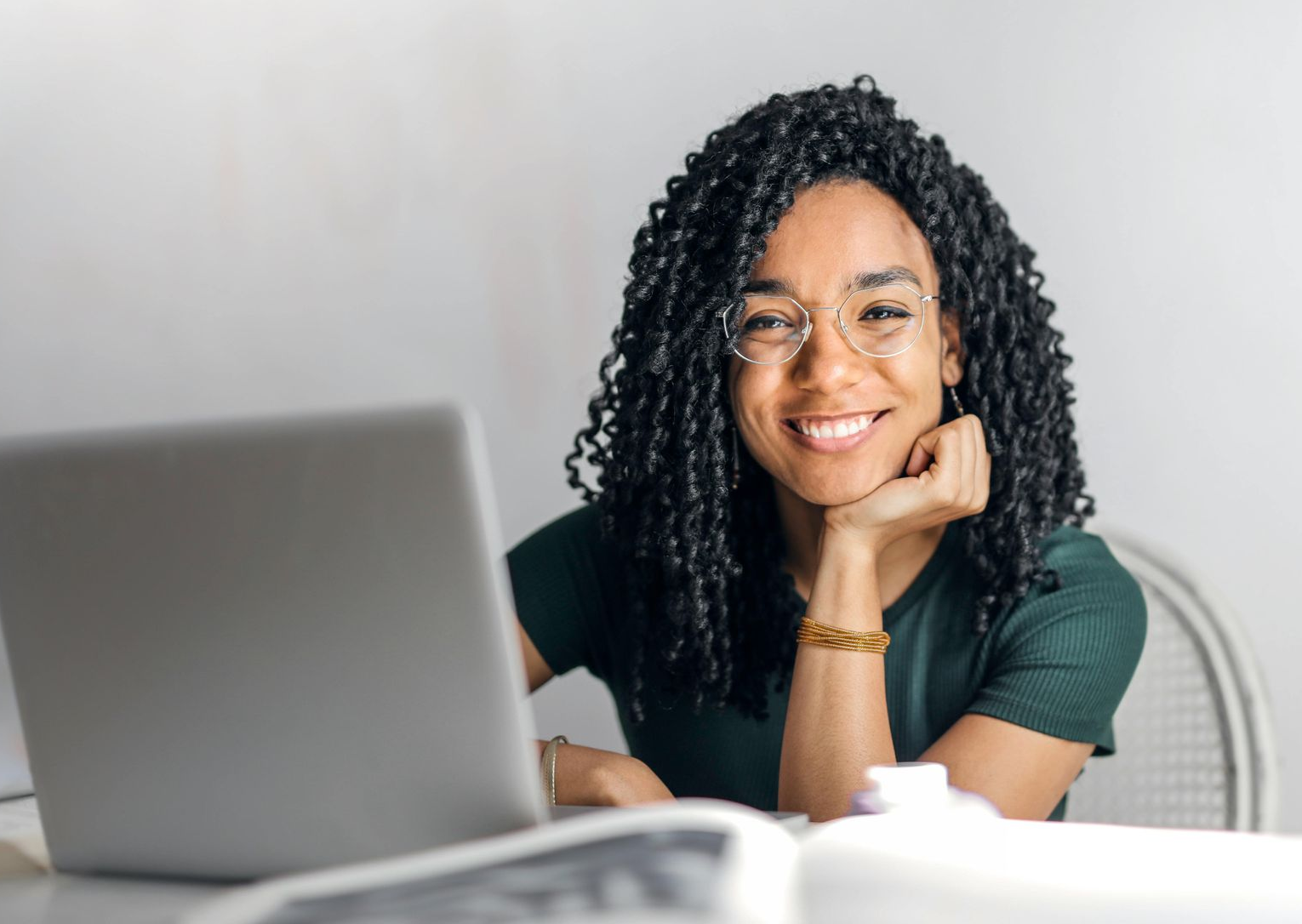 Woman with curly hair and glasses smiles at the camera, sitting at a table with a laptop and book.