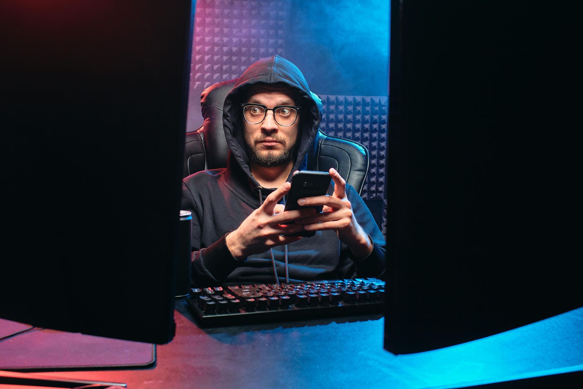 Man in hoodie and glasses, holding phone, sitting at a desk between two monitors. Blue and red lighting.