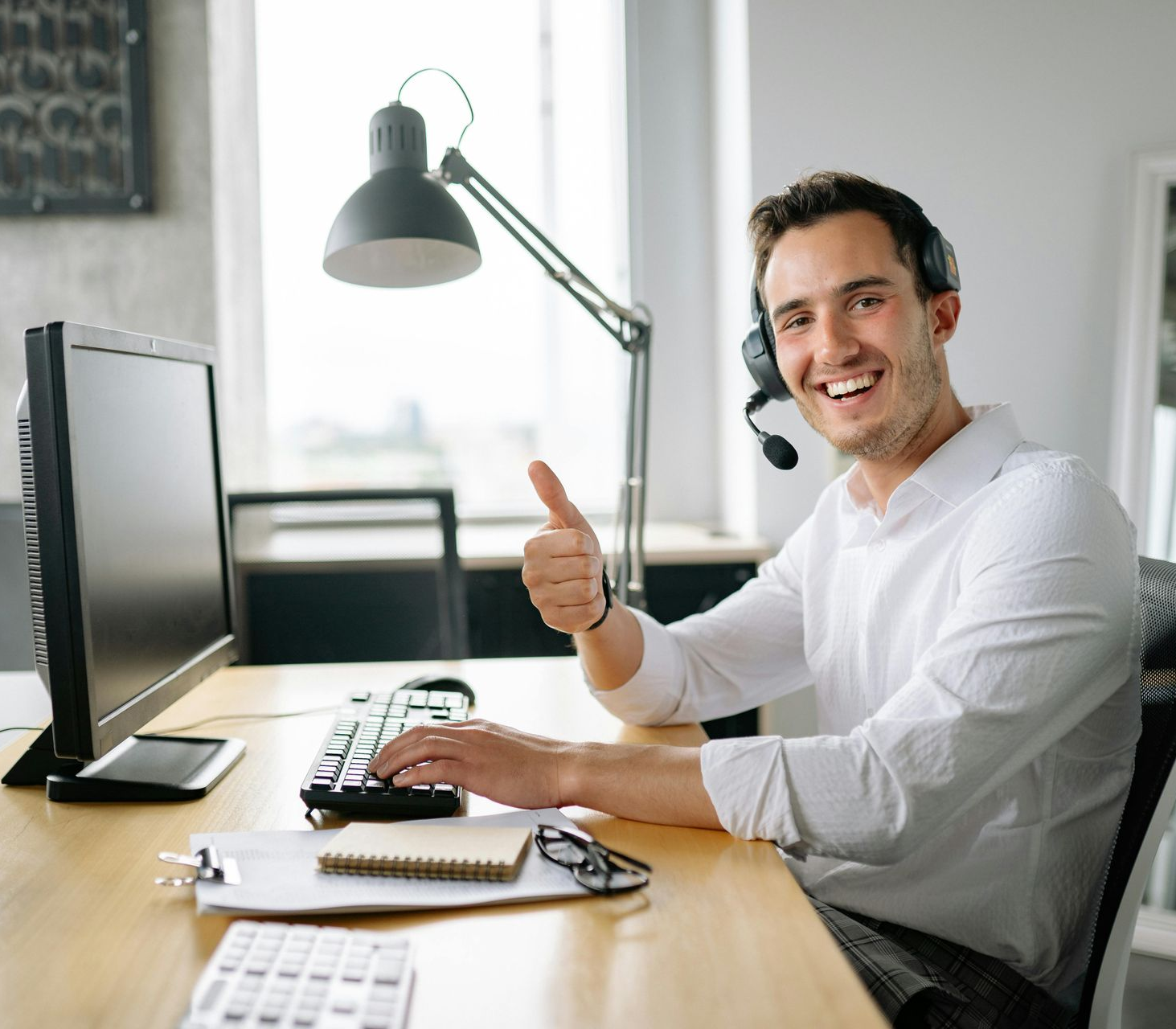 Man in headset at desk with computer, giving thumbs up. Smiling, appears to be in an office setting.