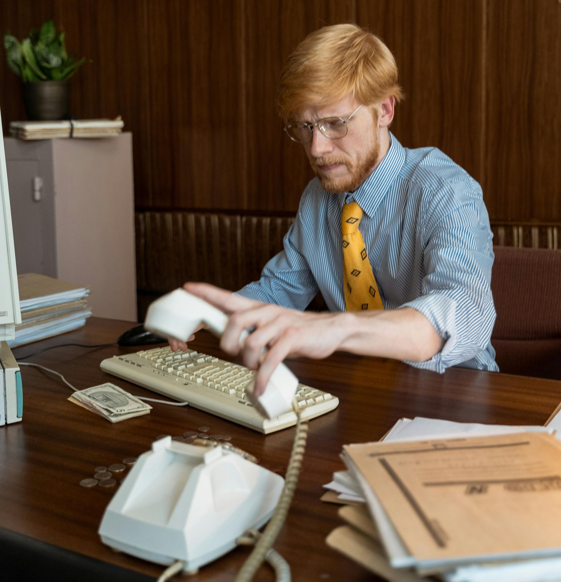 Man with red hair and glasses at a wooden desk holding a phone receiver, looking concerned, vintage office.