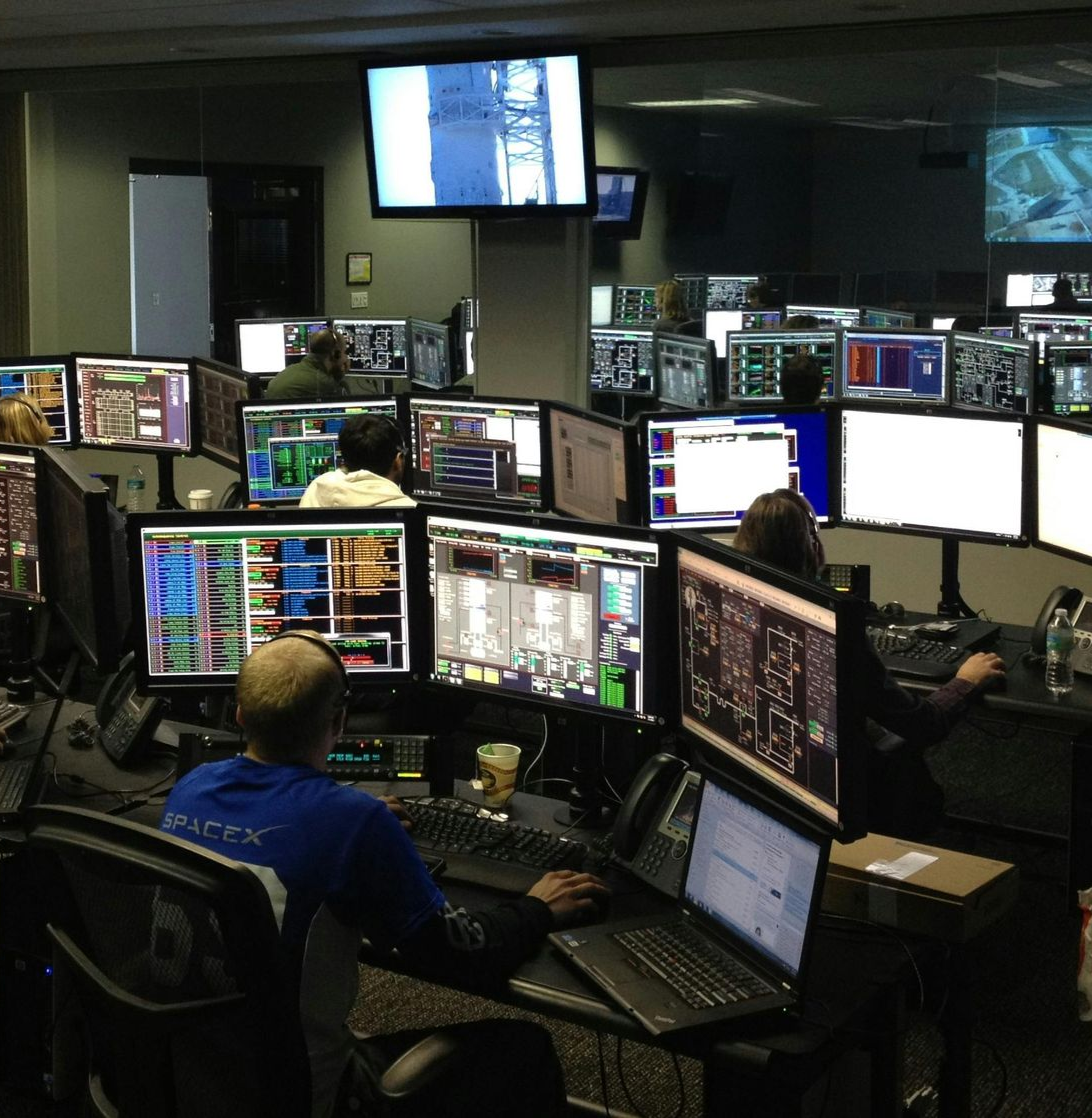 A control room with multiple monitors, operators at workstations, and wall-mounted screens displaying data and visuals.