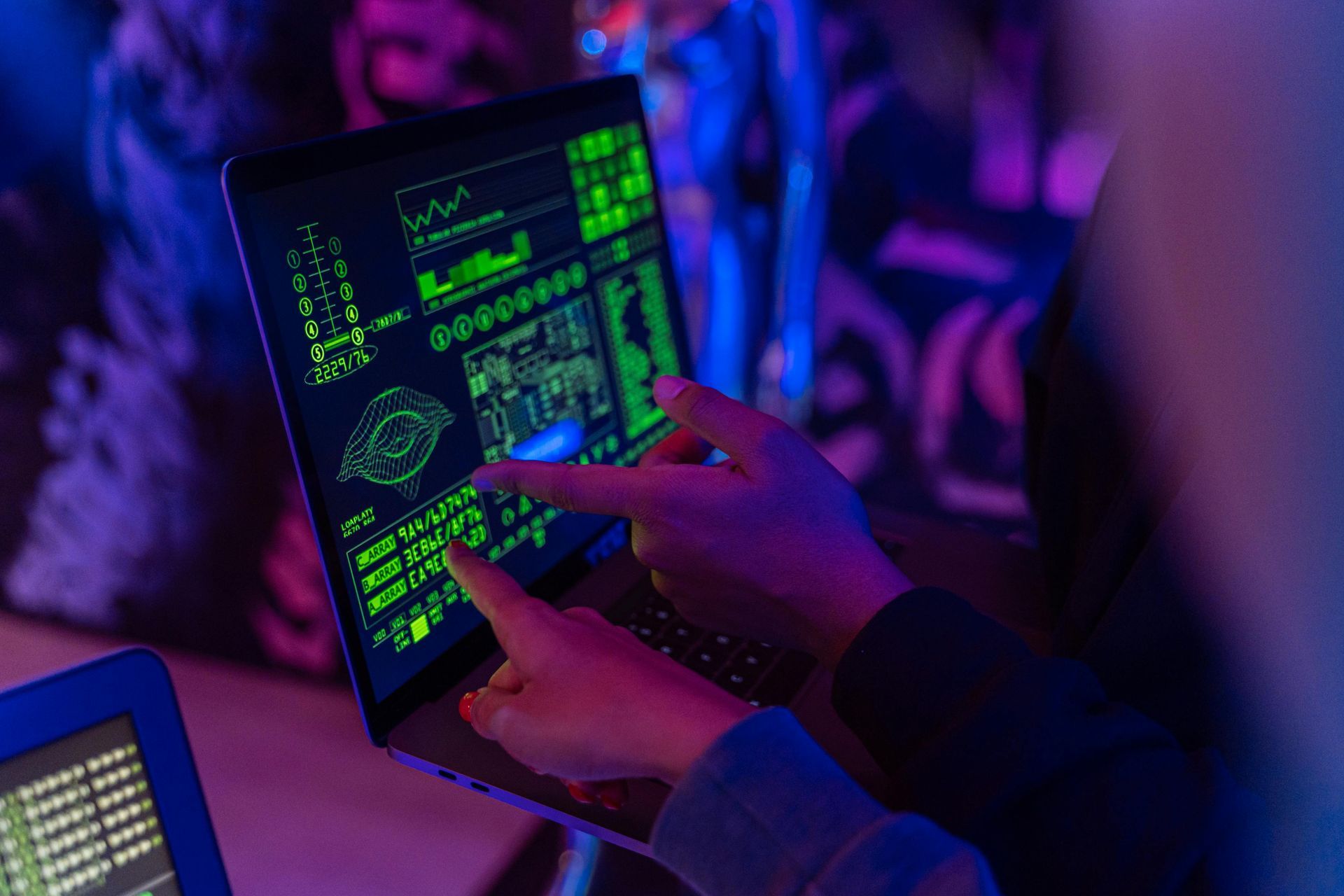 Two hands point at a laptop screen displaying a neon green interface with digital charts and data in a dimly lit room.
