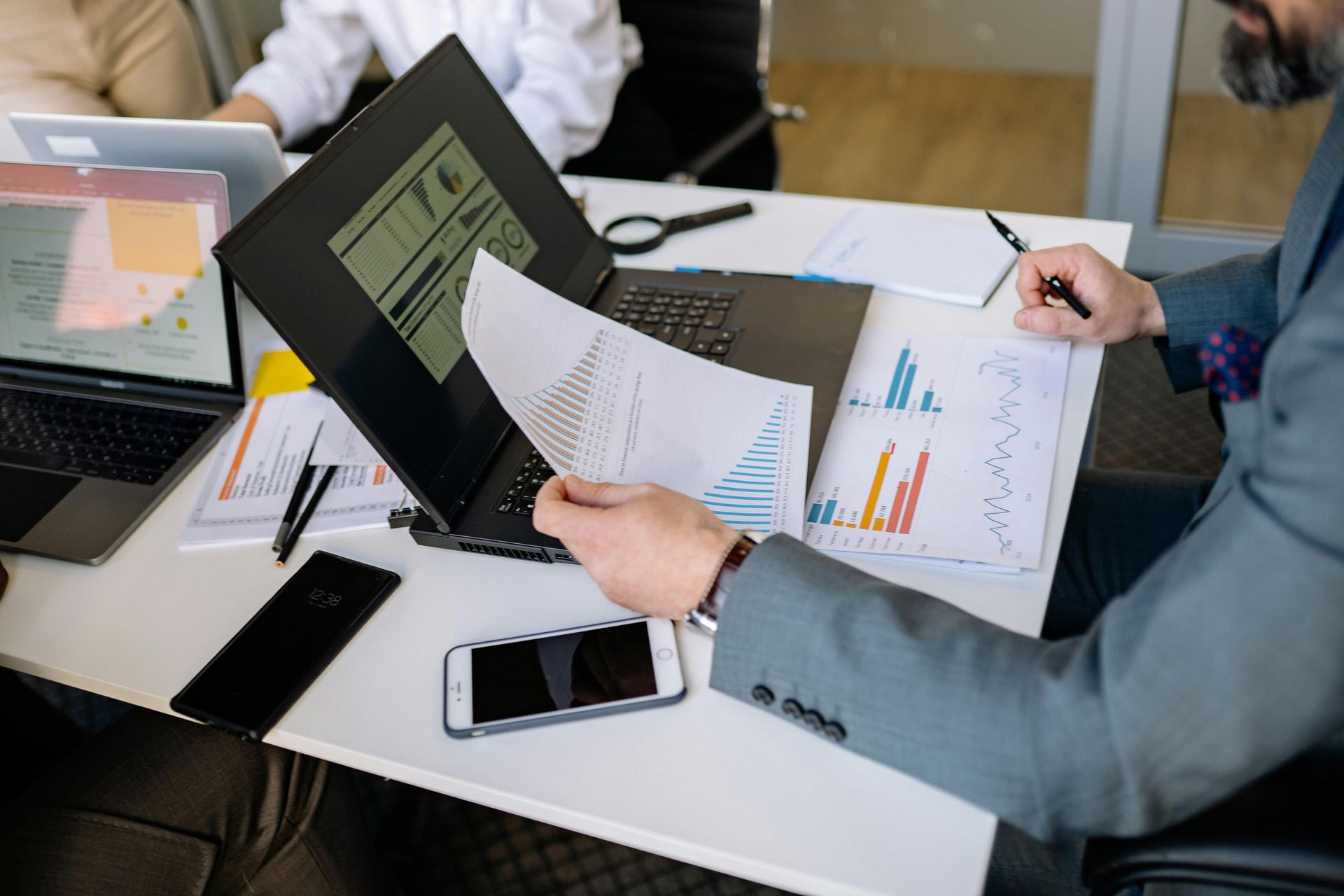 A professional at a desk reviewing charts and data on papers and laptops during a business meeting.