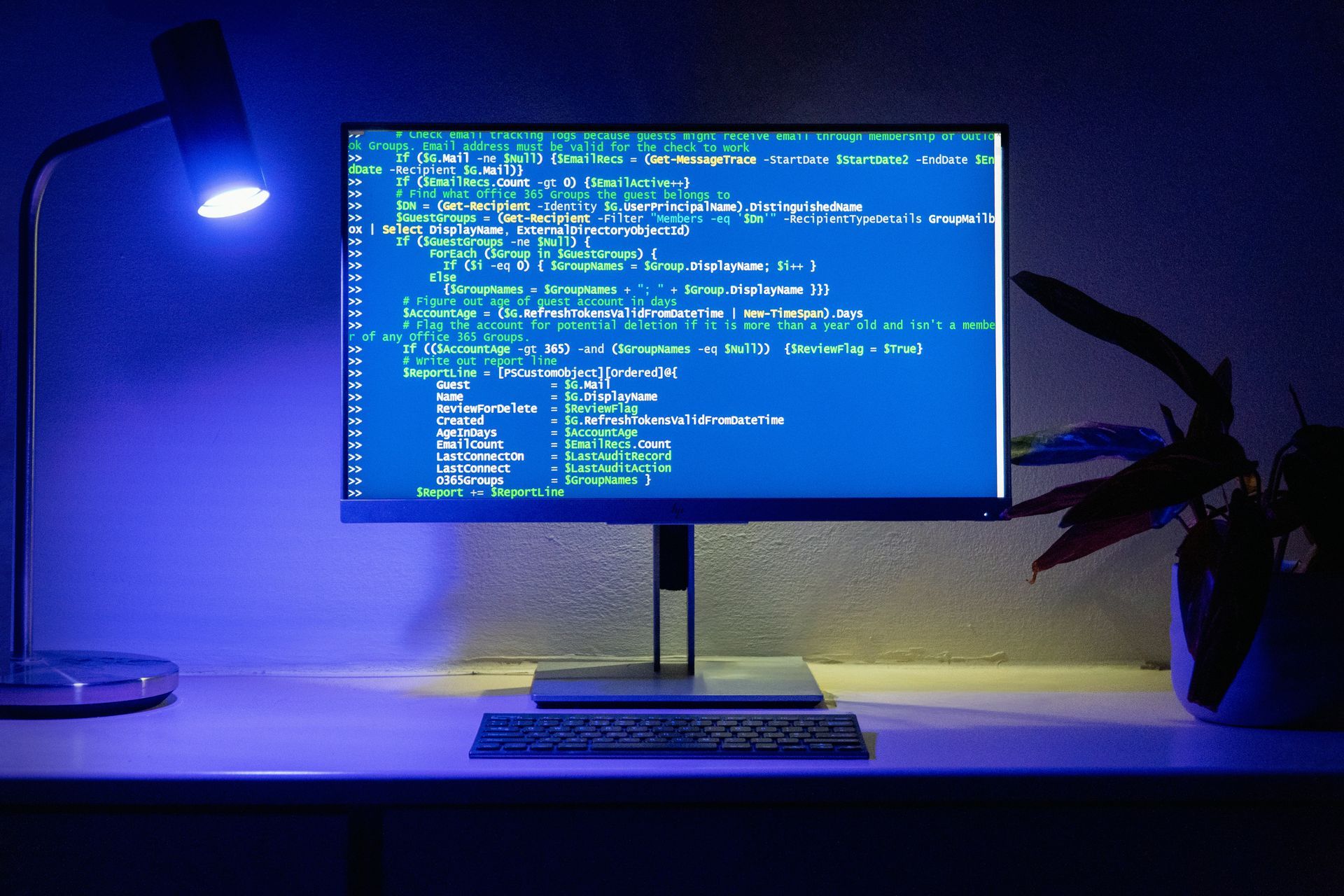 Computer monitor displaying code, illuminated by a blue desk lamp; keyboard visible on a white desk.