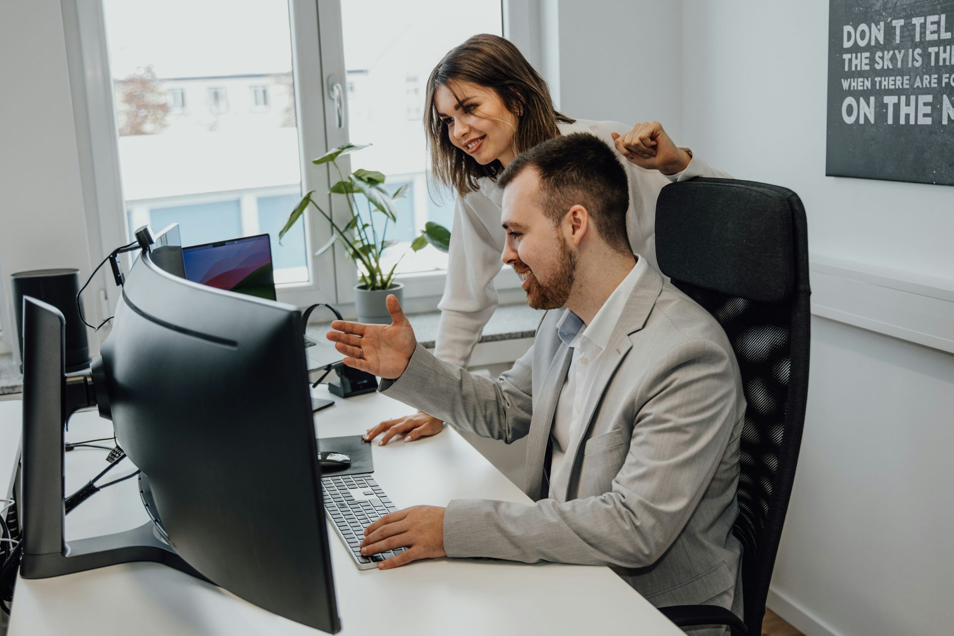 Man and woman at a desk looking at a computer. Woman points while man gestures, both smiling in an office setting.