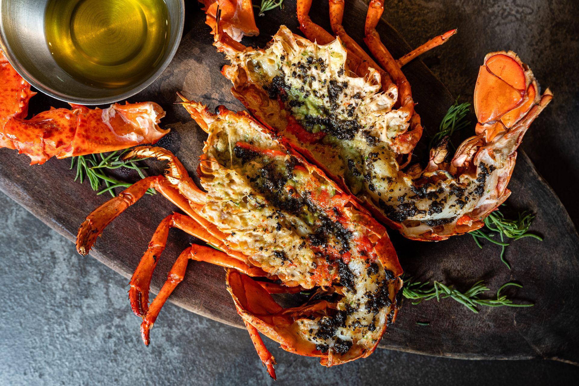 Two lobsters are sitting on a wooden cutting board next to a bowl of oil.