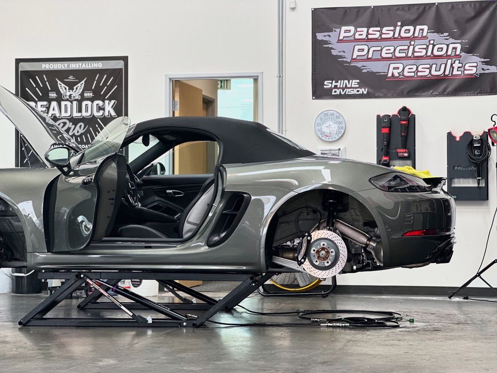 Gray convertible sports car on a lift, door open, wheels removed in a workshop, with banners.