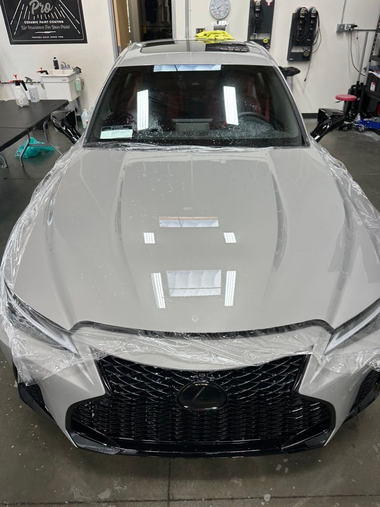 A high-angle view of a grey Lexus sedan inside a garage, with clear plastic film covering its front end and hood.