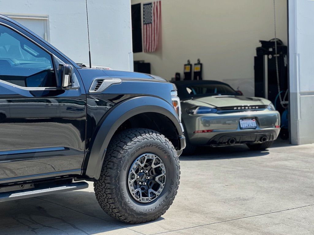 Black truck parked beside a green car in front of a garage. American flag hangs above.