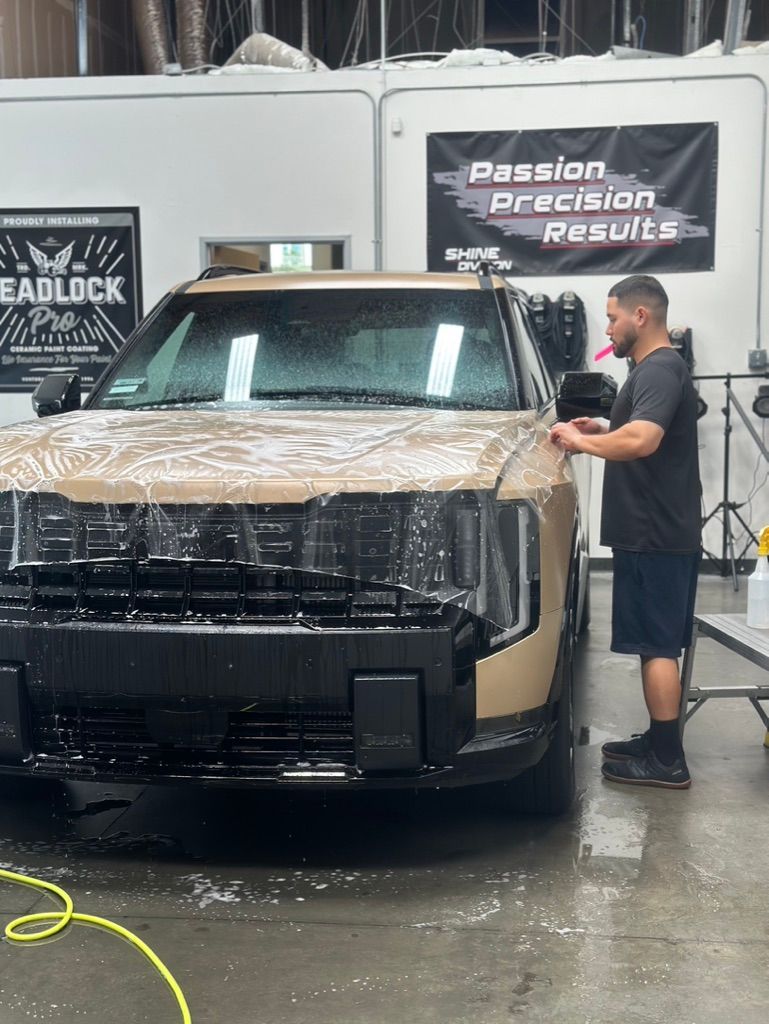 A technician applies a clear paint protection film to the hood of a tan SUV inside a detailing shop.