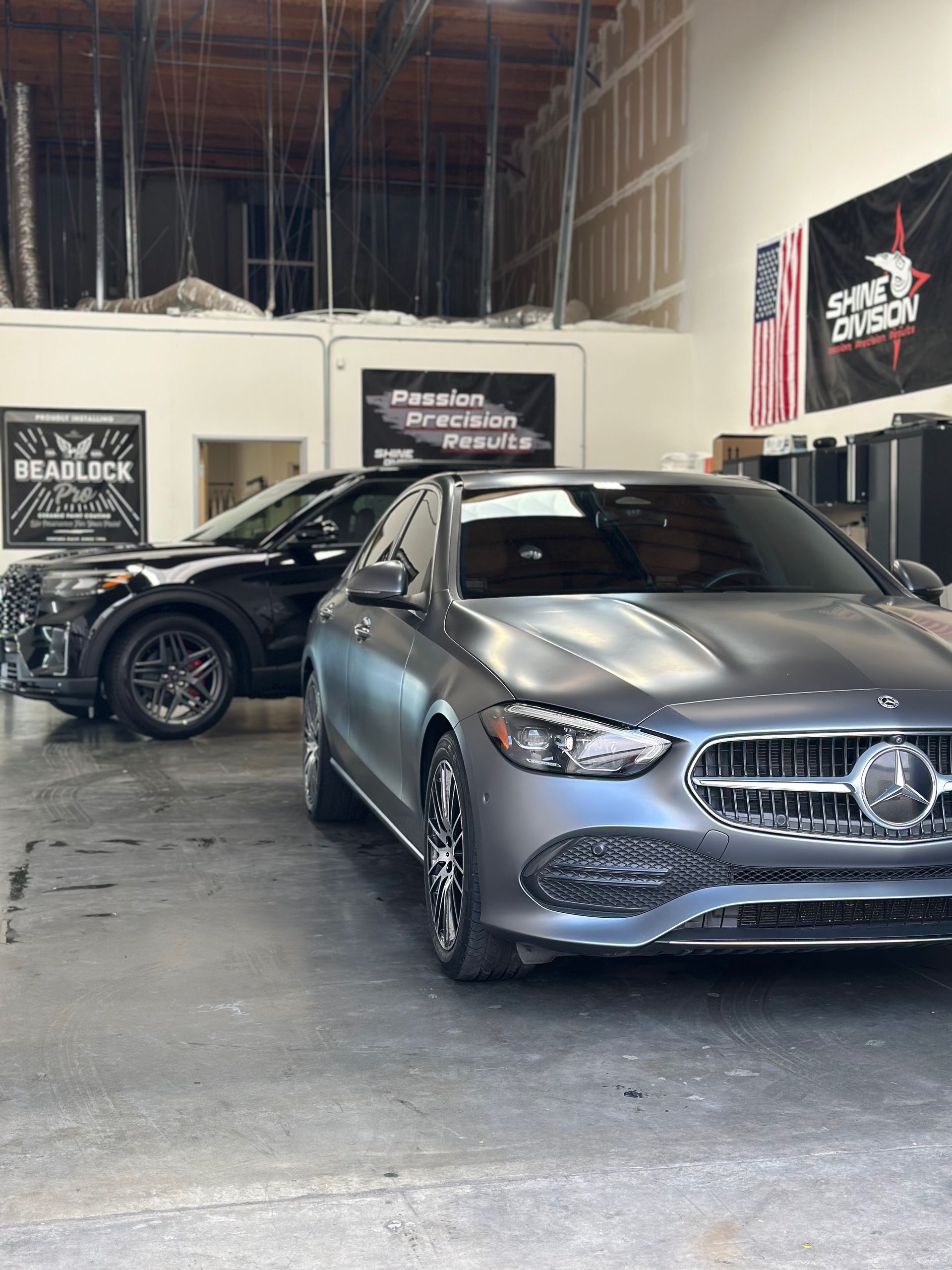 Two cars, a black SUV and a gray sedan, parked inside a garage. American flag and business signs visible.