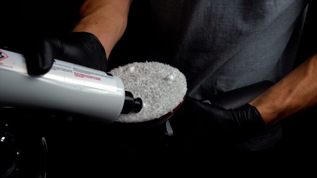 A man wearing black gloves is pouring a bottle of liquid into a polishing pad.