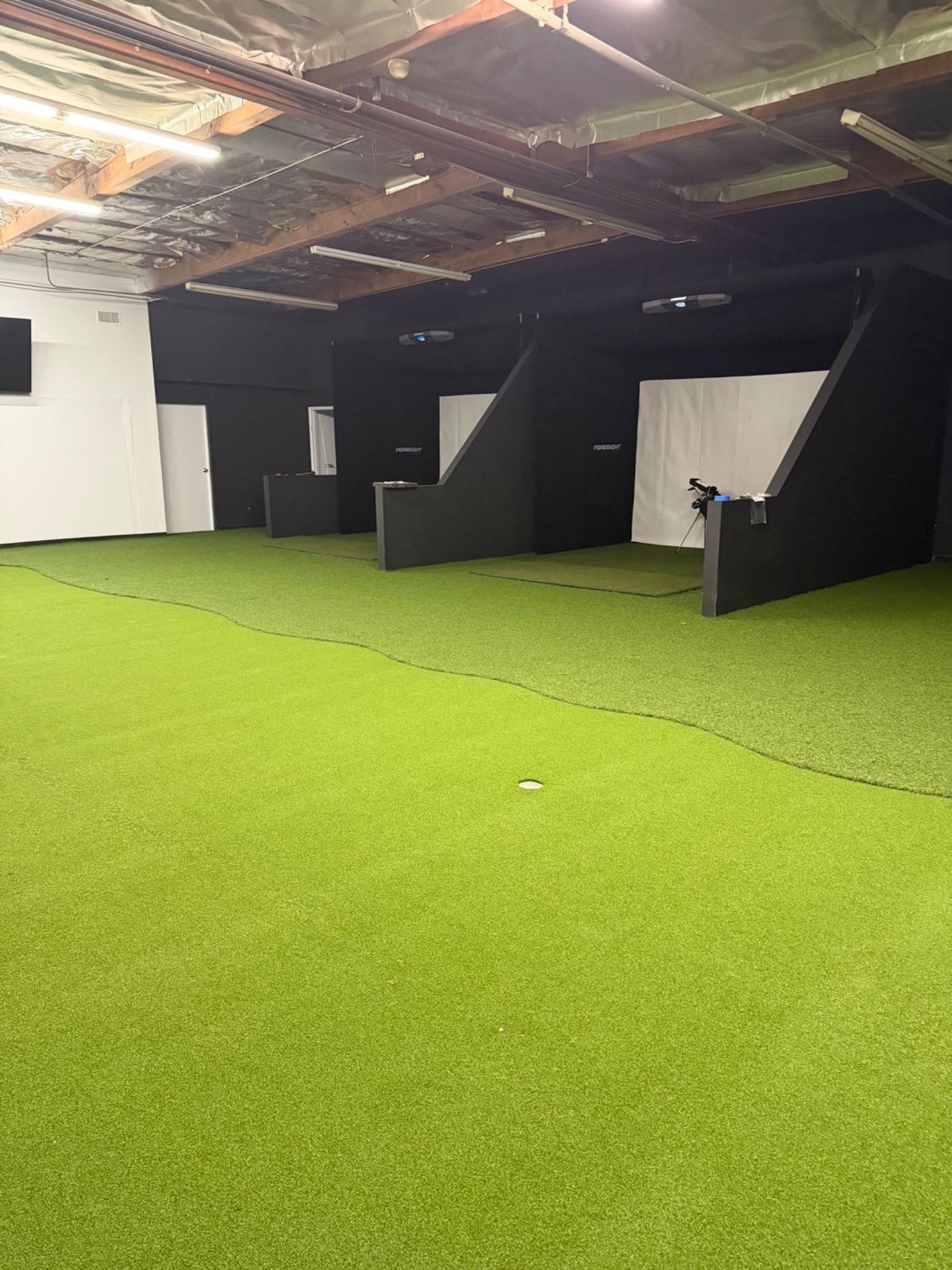 Indoor room with green turf flooring, black cubicle-like structures, and white partition walls under exposed ceiling beams