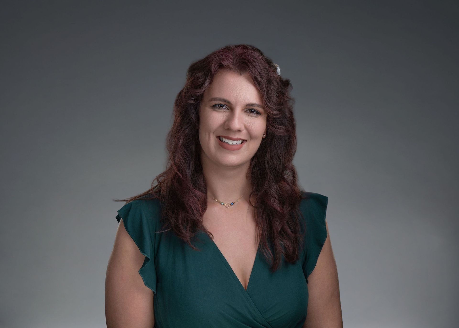 Smiling woman with wavy auburn hair in a teal top against a gray background