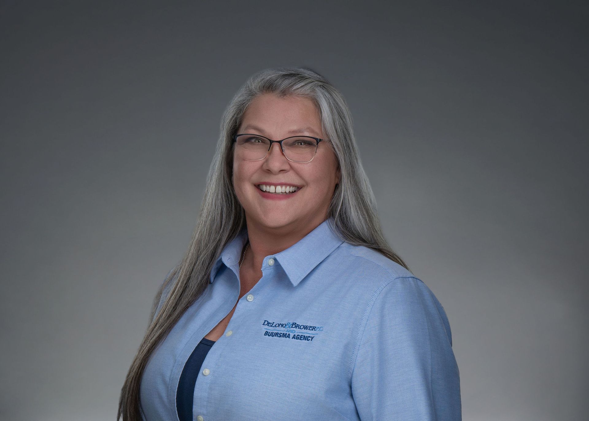 Smiling woman with long gray hair and glasses wearing a light blue shirt against a gray background