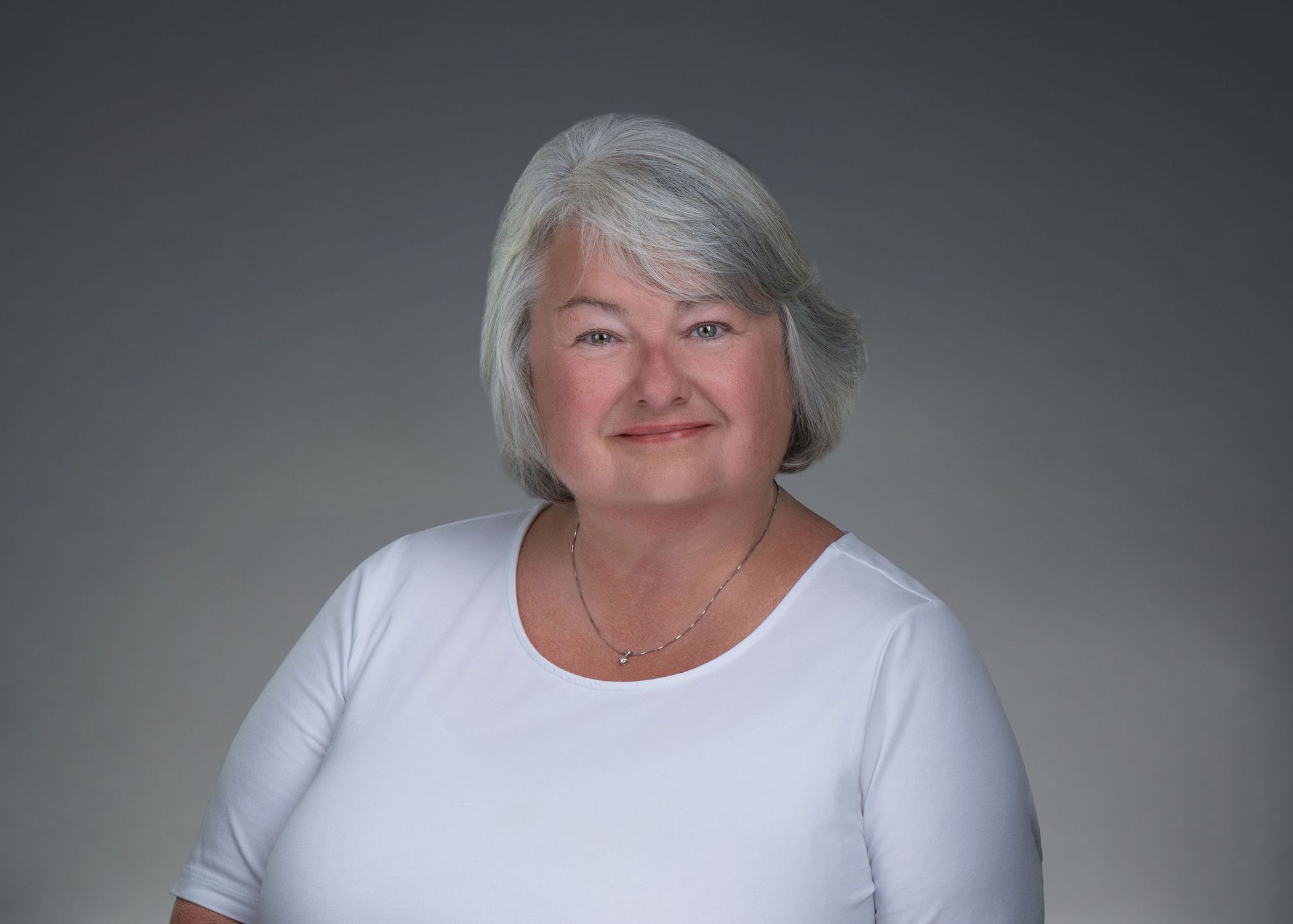 Smiling woman with short gray hair wearing a white top against a gray background