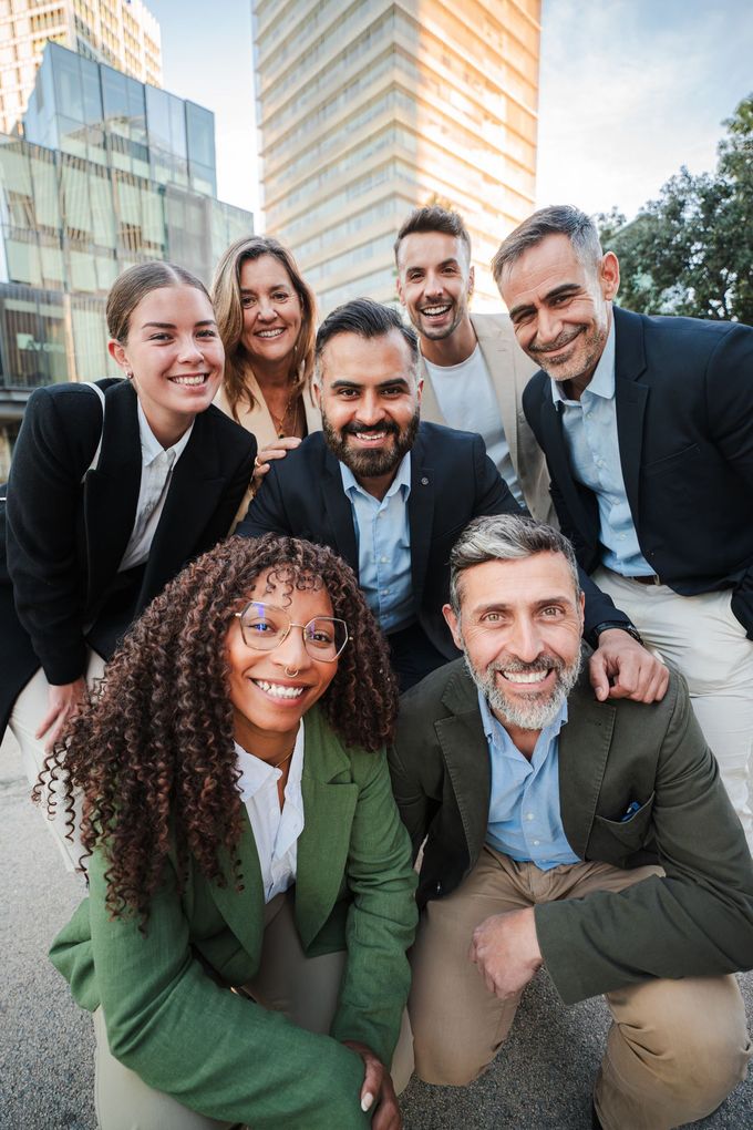 Seven coworkers smiling outdoors in business attire in front of office buildings