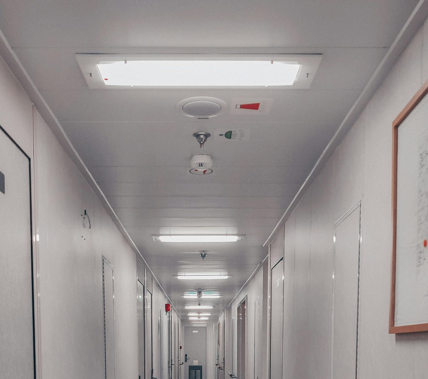 Hallway with white walls and ceiling, doors, lights, and a framed drawing on the right.