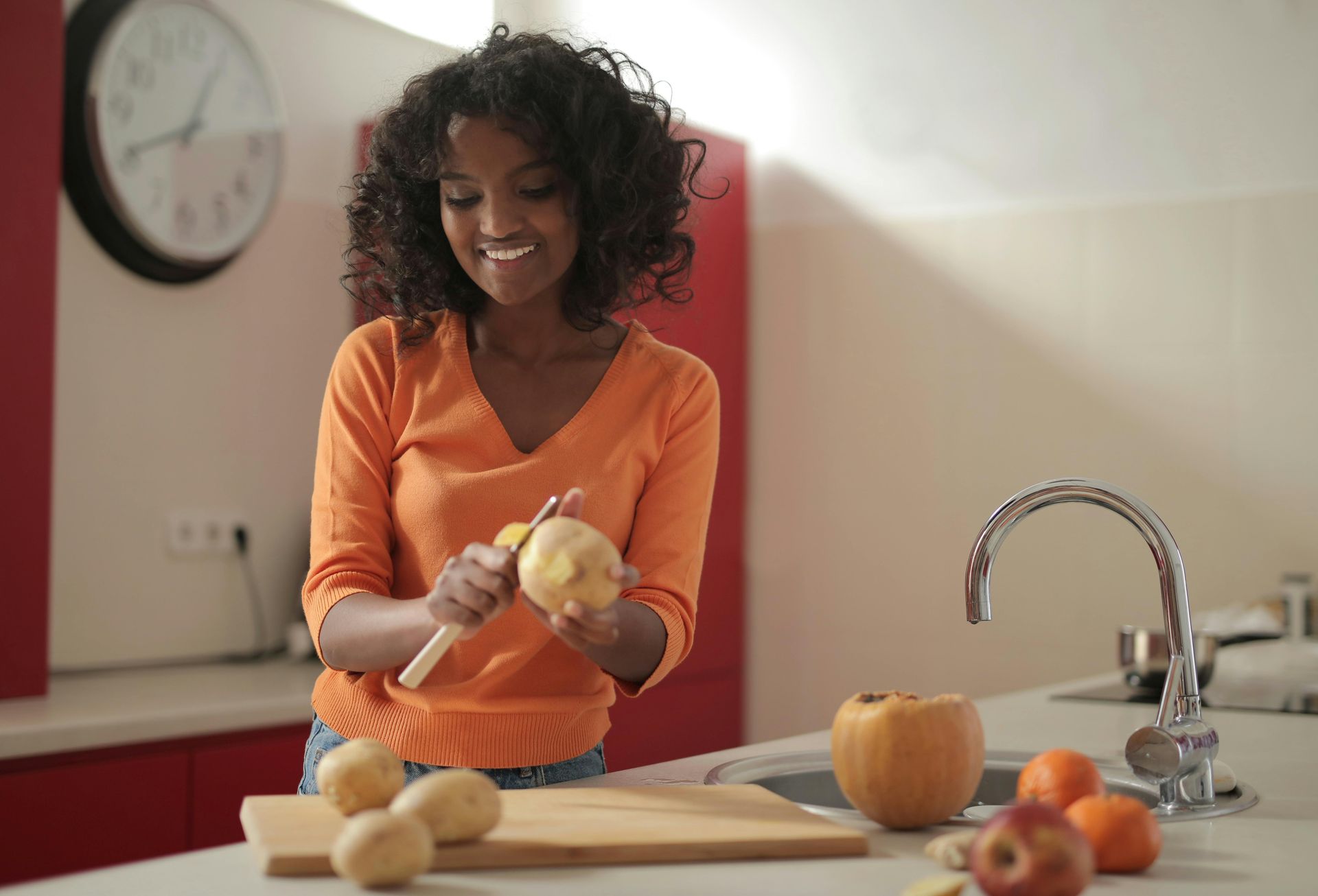 Woman peeling potato in kitchen, smiling. Bright orange sweater.
