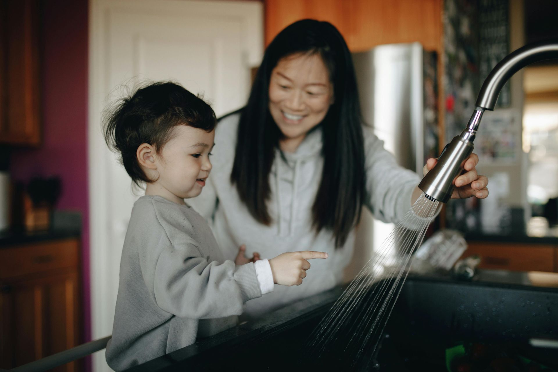 Woman in red cardigan slices tomatoes, child watches at a table in the kitchen.