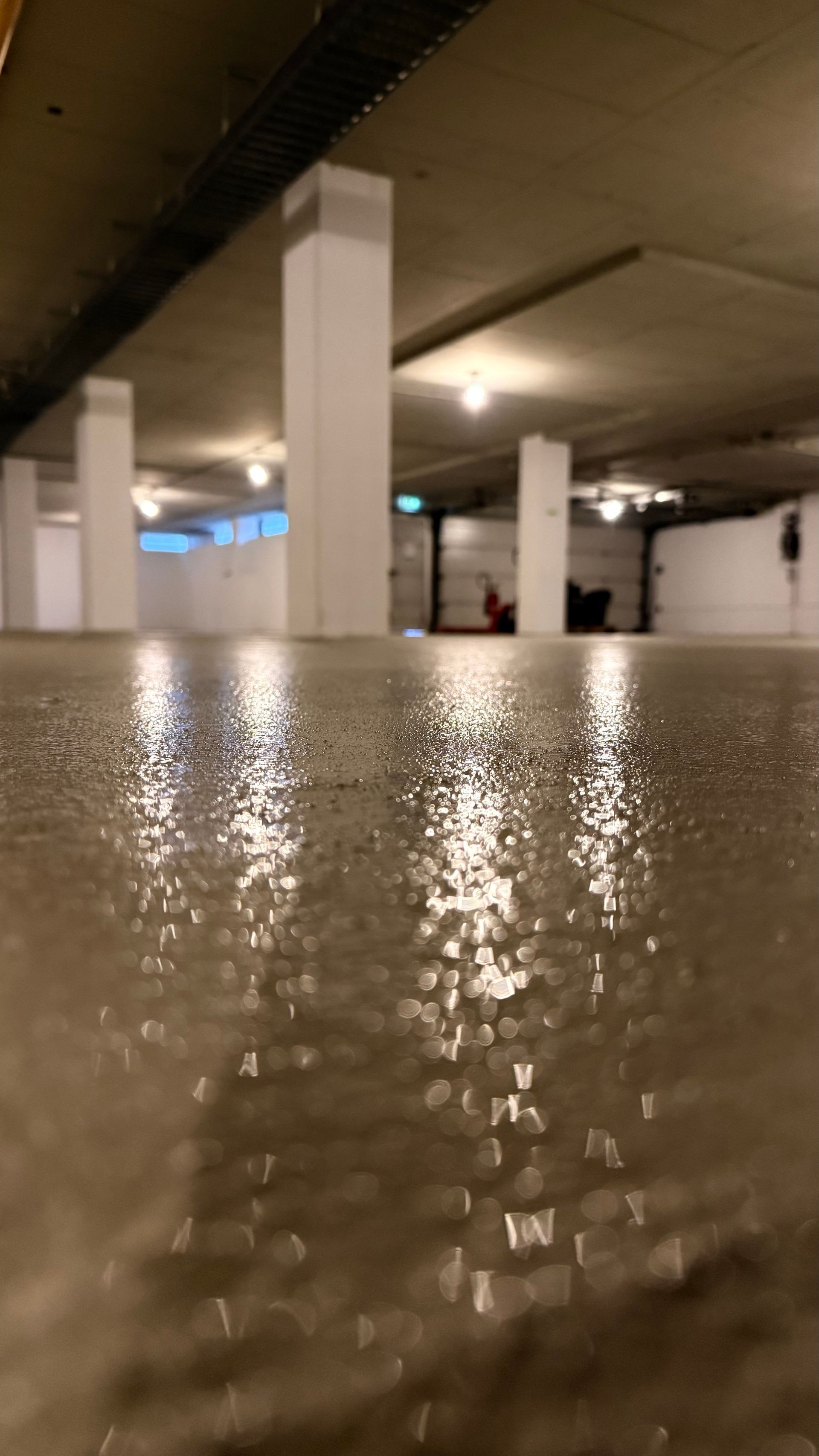 Een natte, reflecterende vloer in een parkeergarage met betonnen pilaren en plafondverlichting.