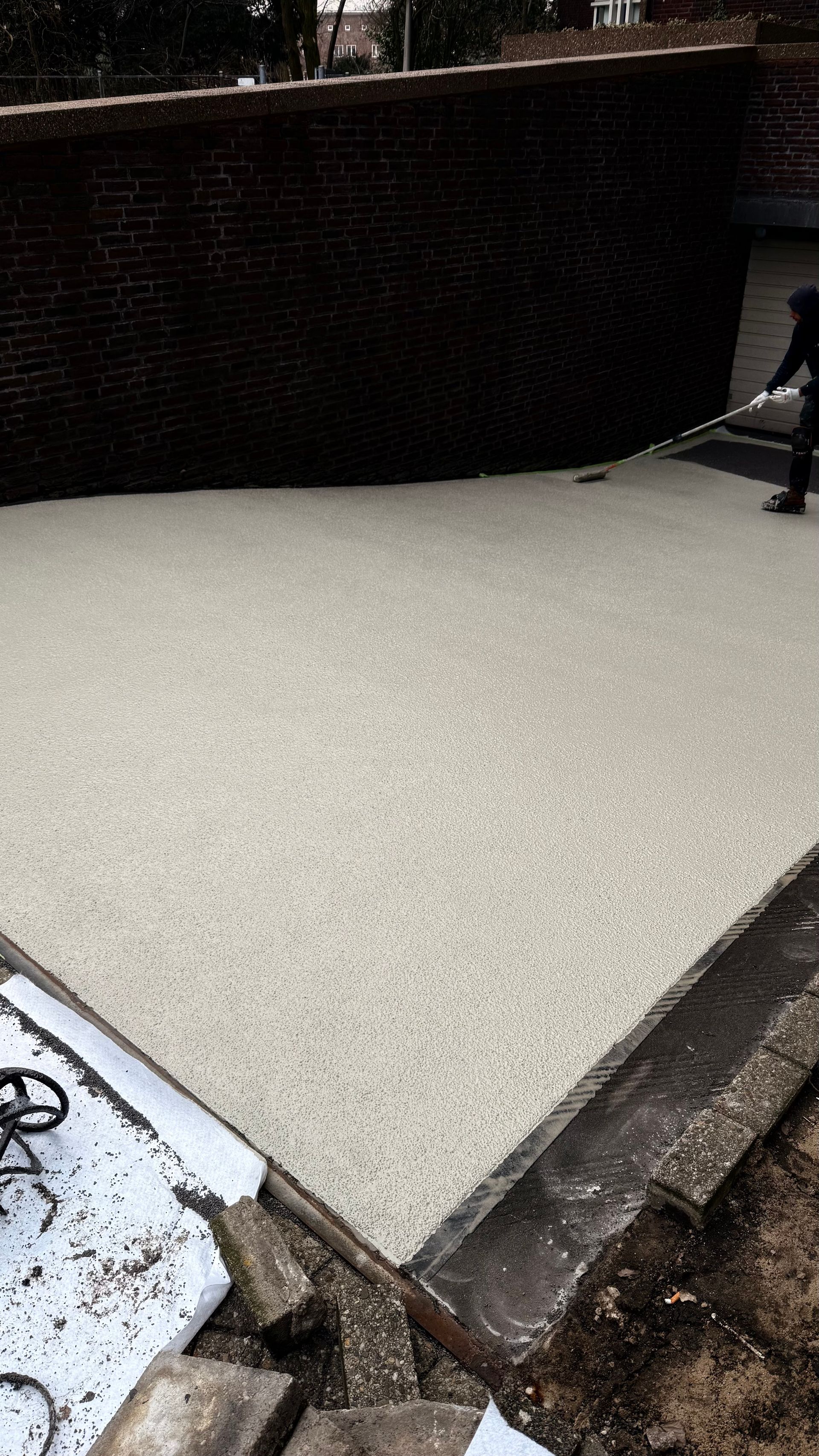 Een persoon brengt een gespikkelde coating aan op een betonnen terras naast een bakstenen muur.