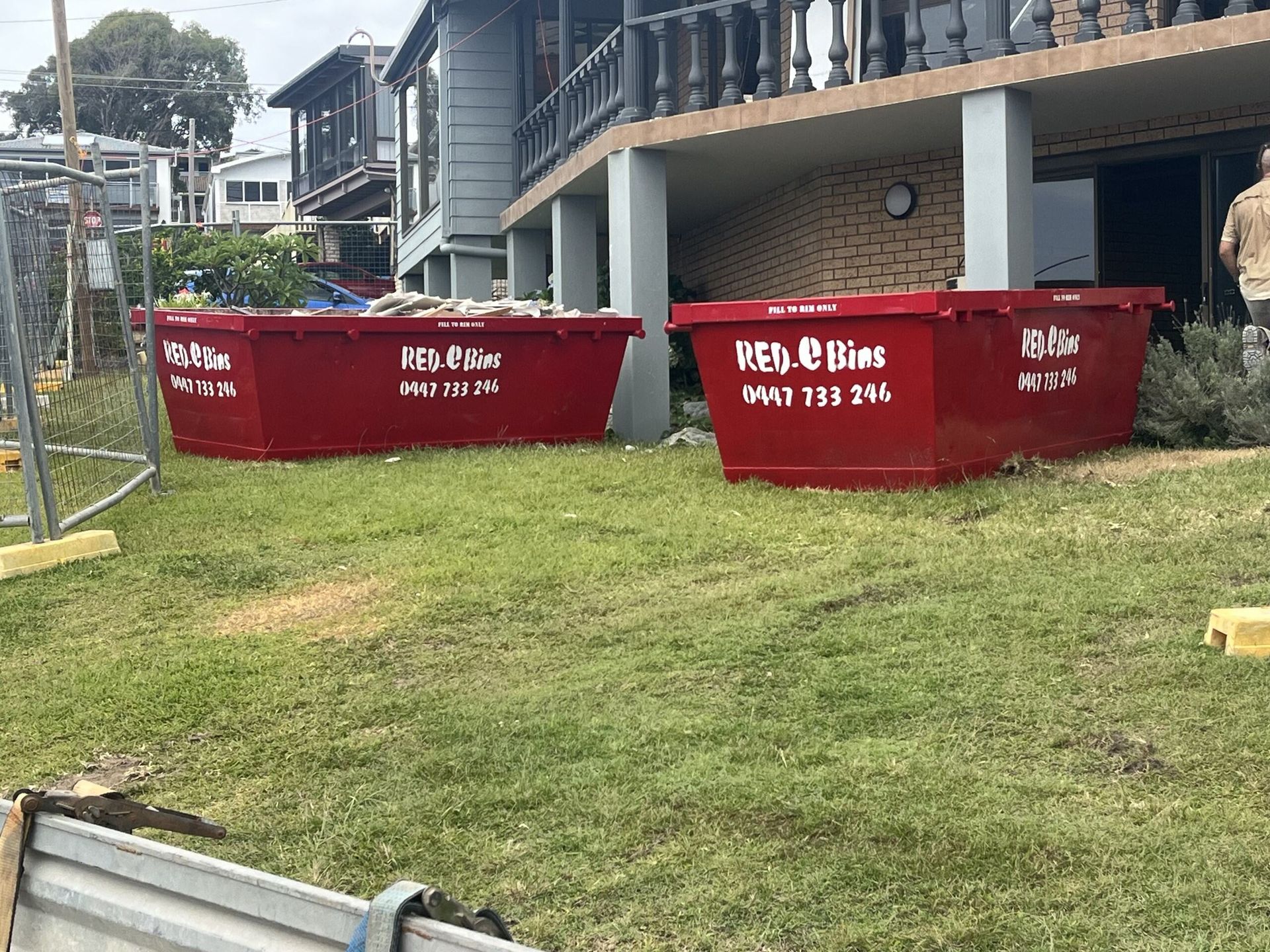 Two red skip bins are sitting in the grass in front of a house.  — Red E Bins In Lake Cathie, NSW