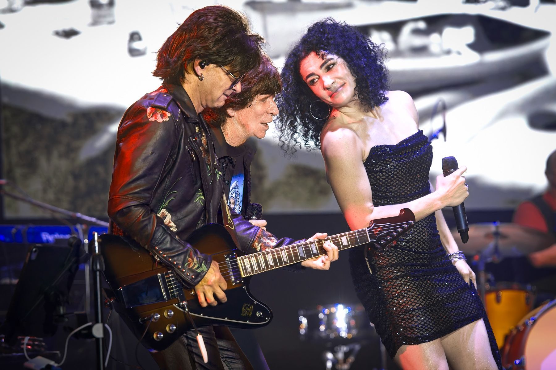 Two musicians performing onstage, one playing guitar while a singer dances beside him.