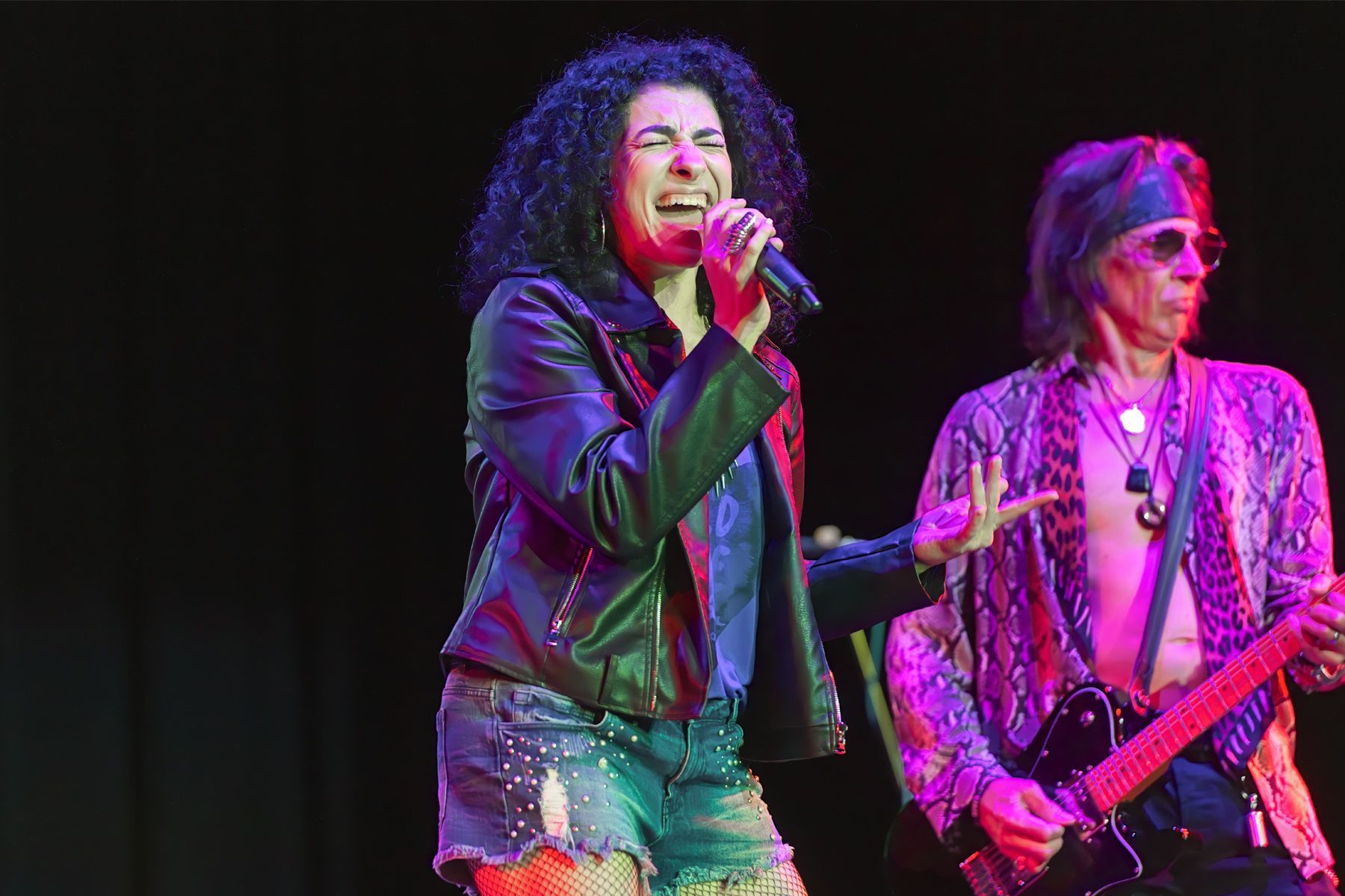 Singer in black jacket performs onstage beside a bassist under pink and purple lights.
