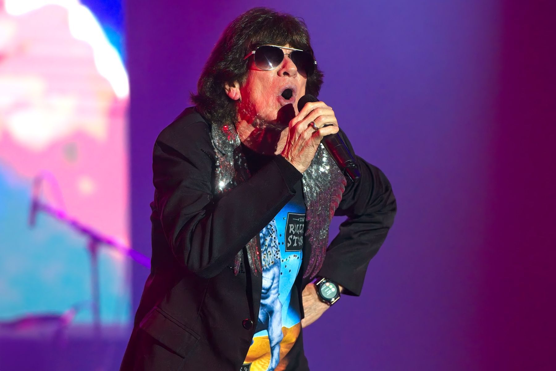 Singer in sunglasses and black jacket singing on a colorful stage with a microphone
