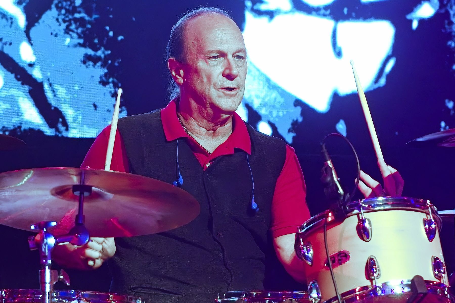 Drummer in red shirt and black vest playing a drum kit onstage under blue and purple lights
