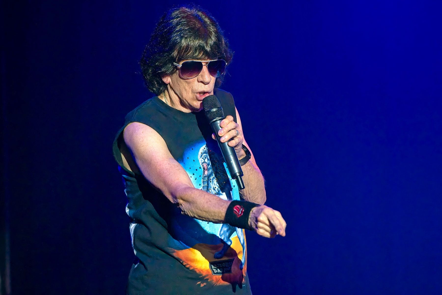 Singer on stage wearing sunglasses and a sleeveless shirt, holding a microphone under blue-purple lighting