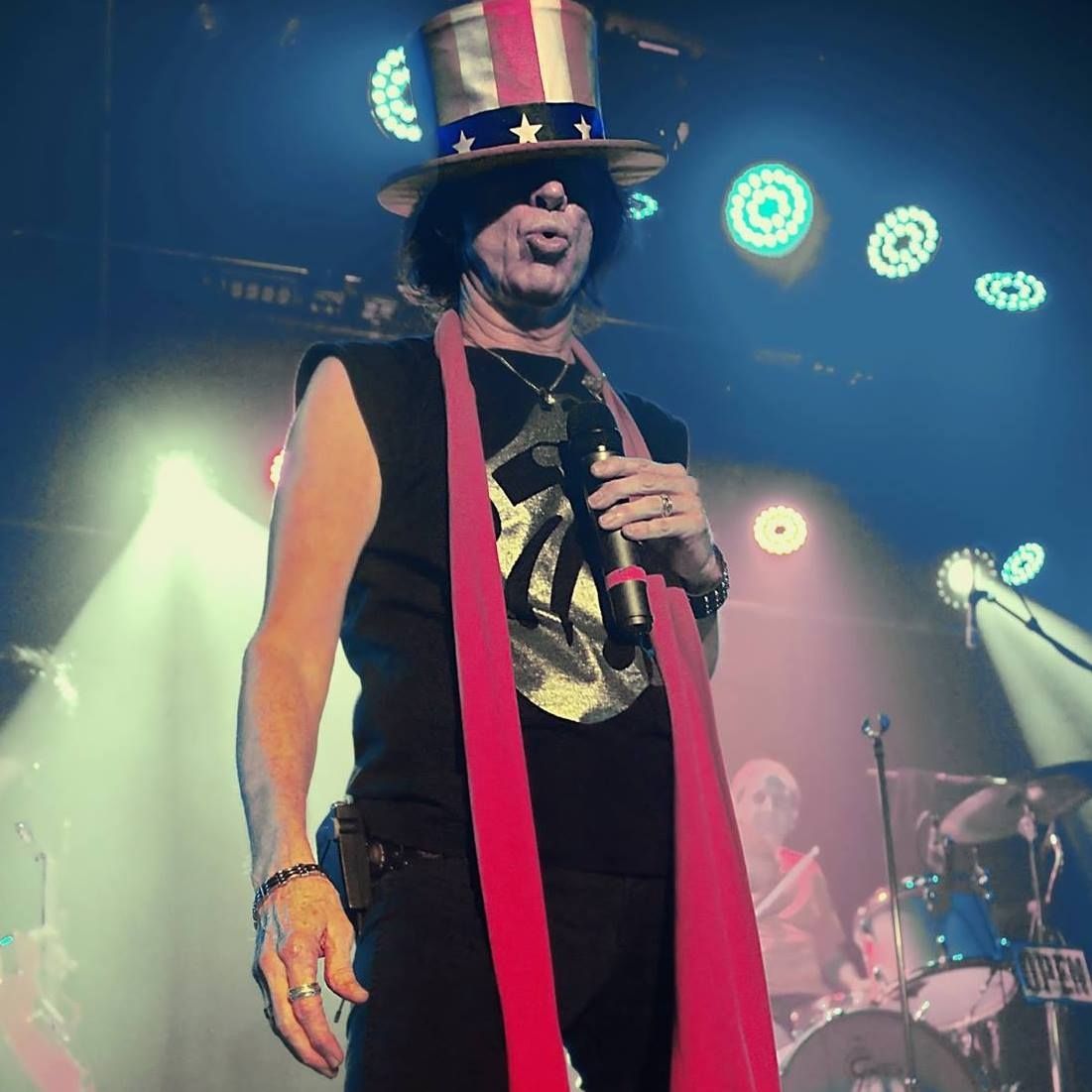 Singer on stage wearing a star-spangled top hat and red scarf under colorful concert lights