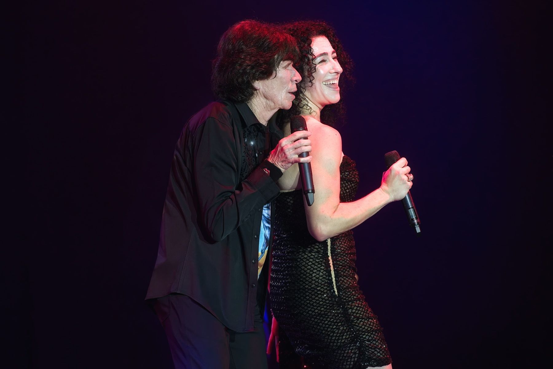Two performers singing on a dark stage, one in black and one in a sparkly dress, smiling and holding microphones.