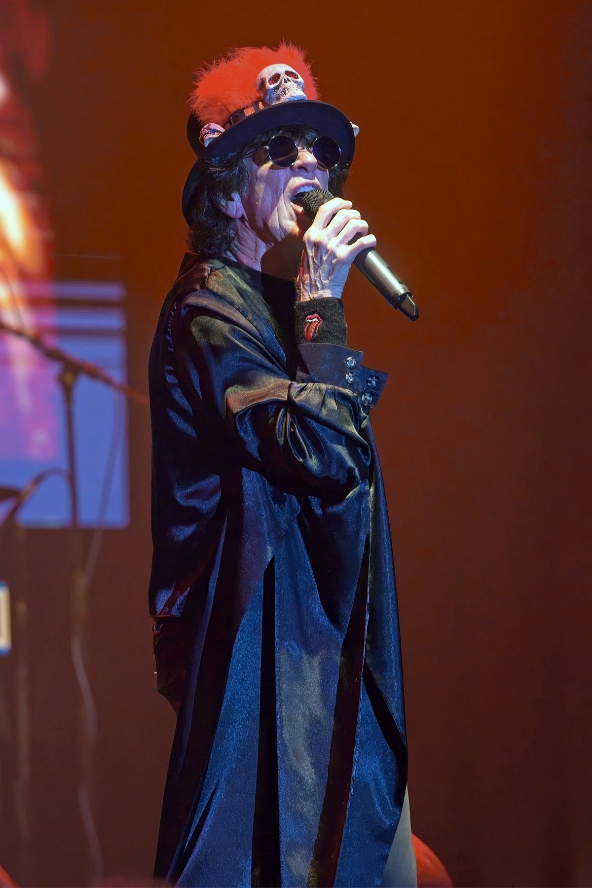 Singer in a black coat and hat, holding a microphone on a red-lit stage.