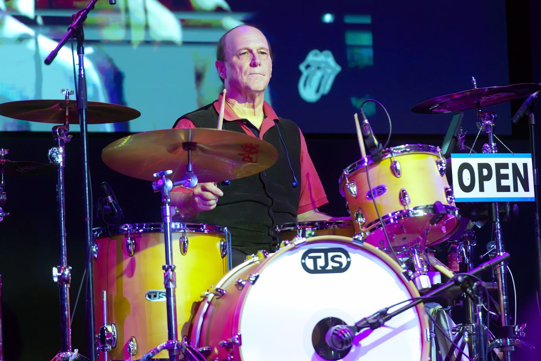 Drummer performing on a brightly lit stage with yellow drums and a visible “OPEN” sign behind him