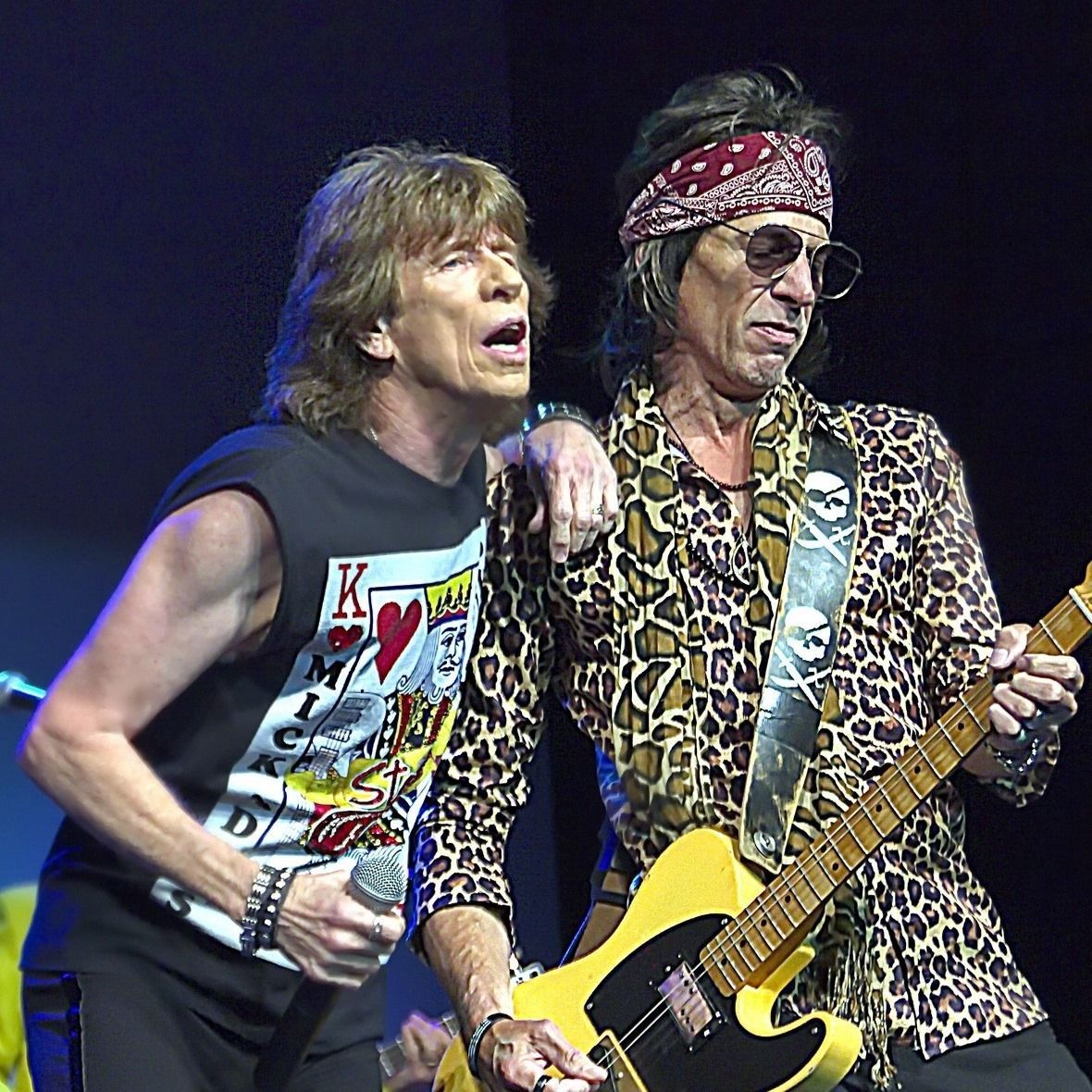 Two rock musicians performing on stage, one singing and one playing yellow guitar, under dark lighting