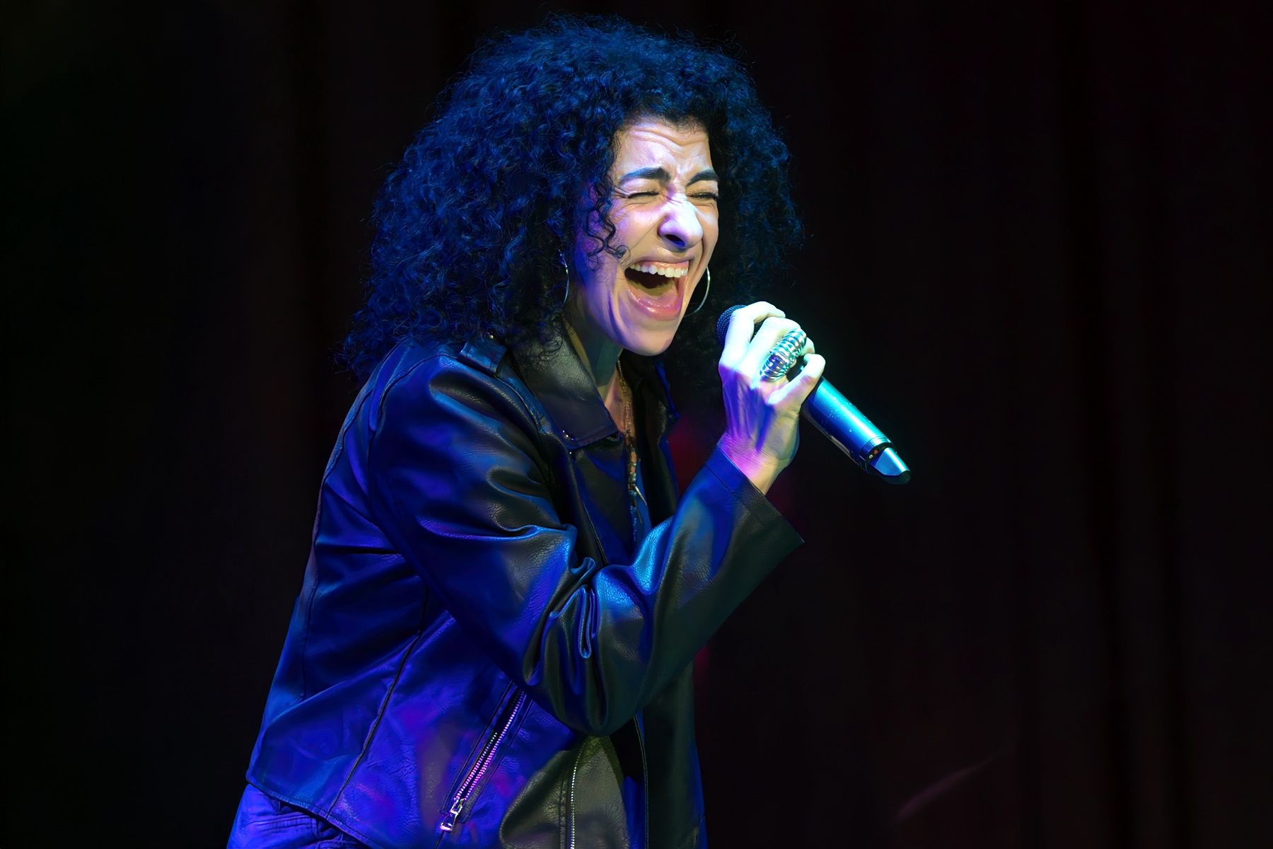 Singer performing on stage, holding a microphone, with blue lighting and a black leather jacket.
