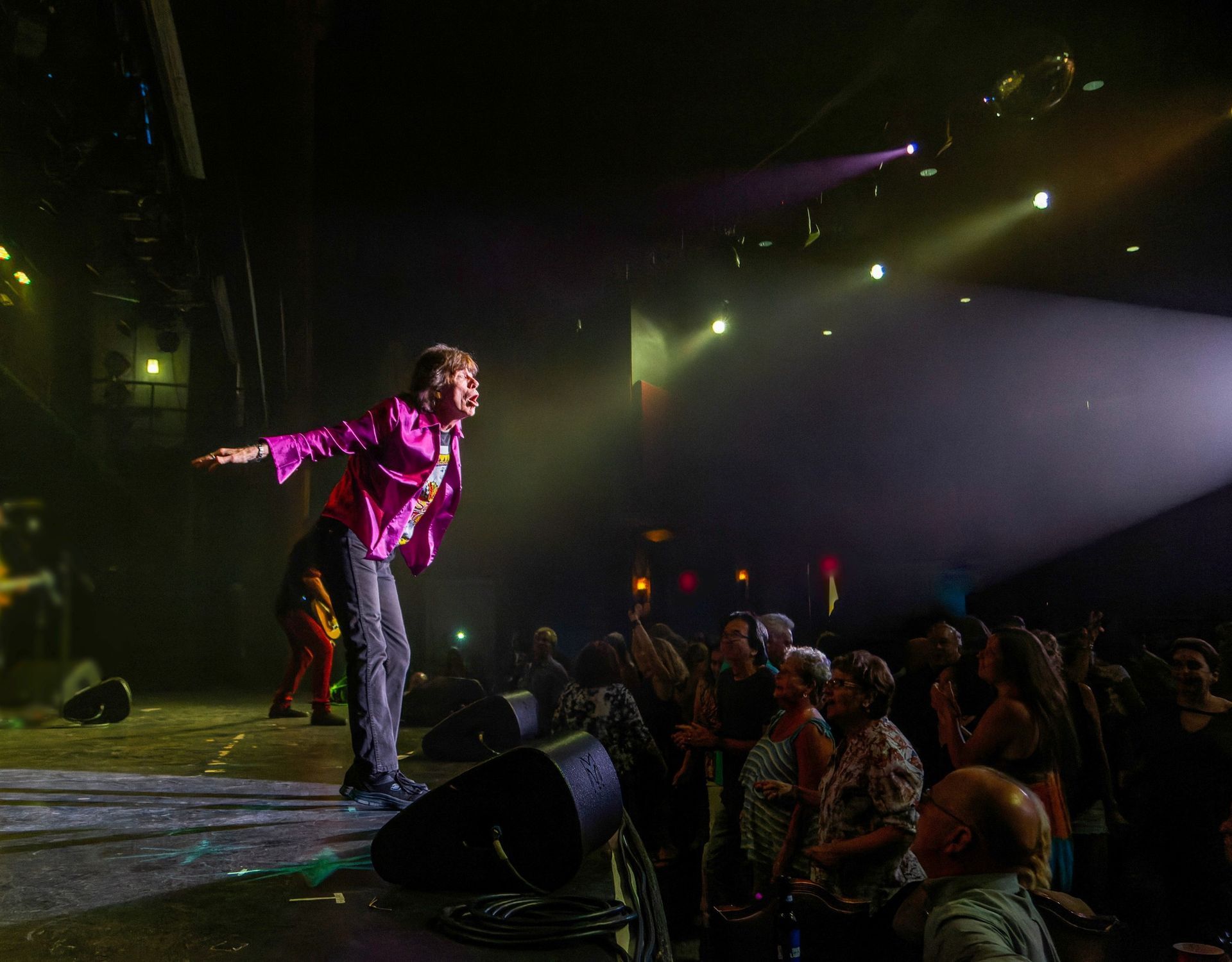 Singer in a bright pink jacket performing onstage, leaning toward an audience under spotlights.