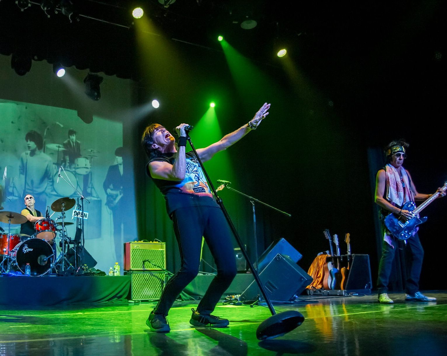 Rock band performing on stage under green lights, singer leaning back at center with drums and guitar behind.