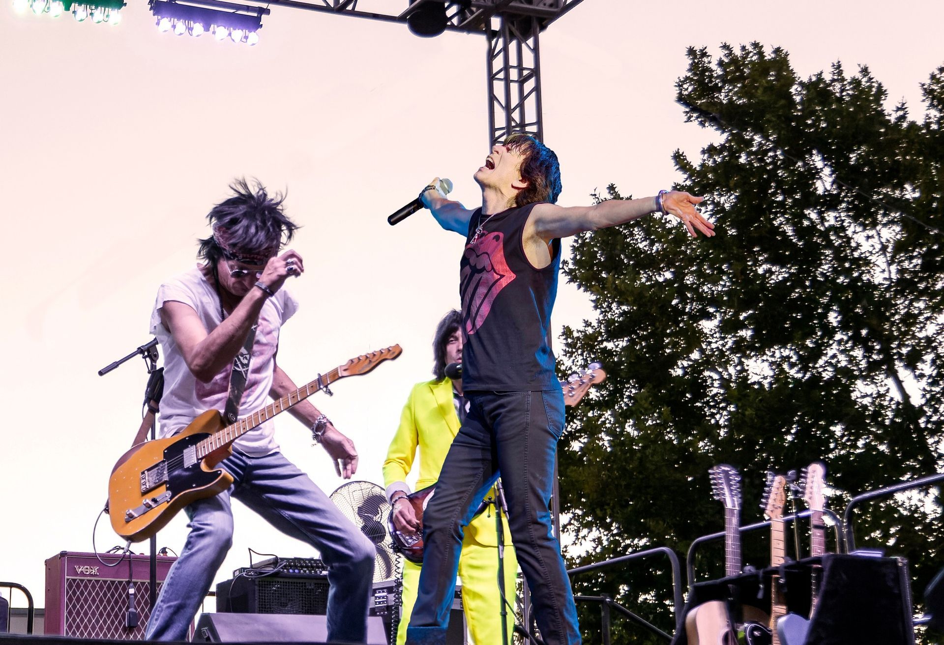 Live band performing outdoors onstage, singer with arms outstretched and guitarist leaning into the music at sunset.