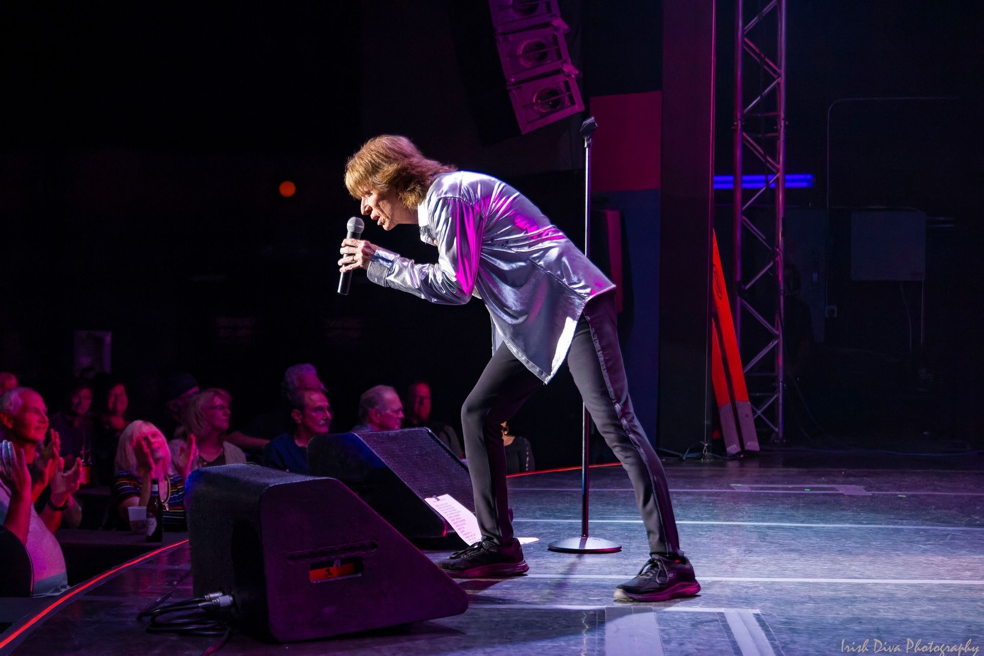 Singer performing on stage under purple lights, leaning toward the audience with a microphone.