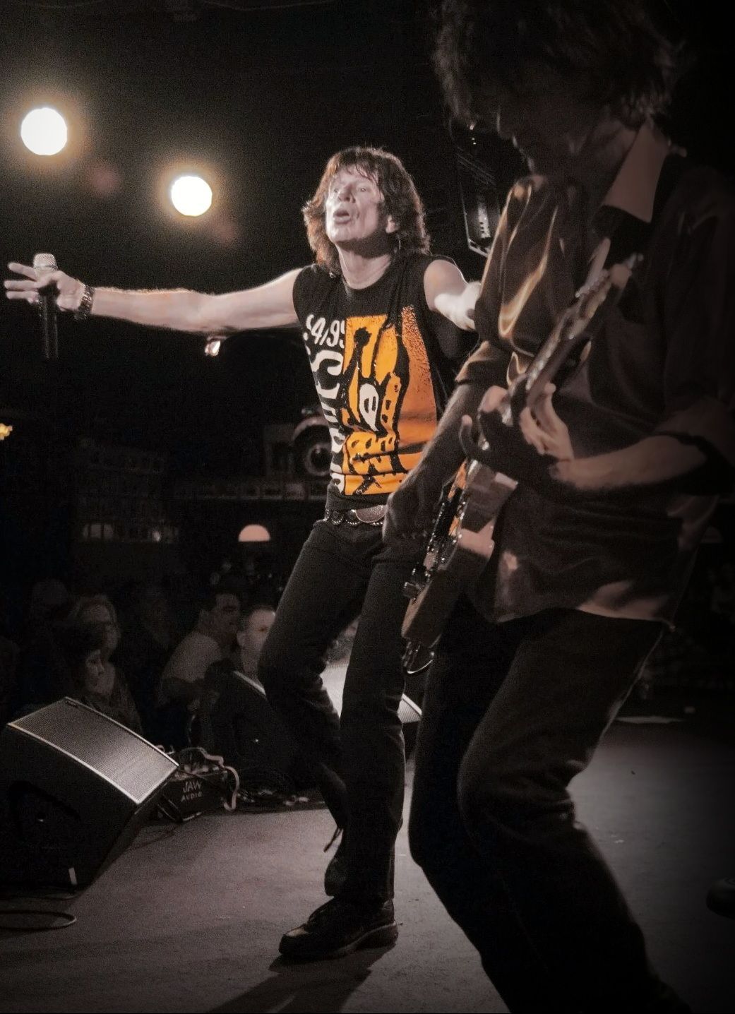 Live rock performance with two musicians on stage, one singing with arms outstretched under bright lights.