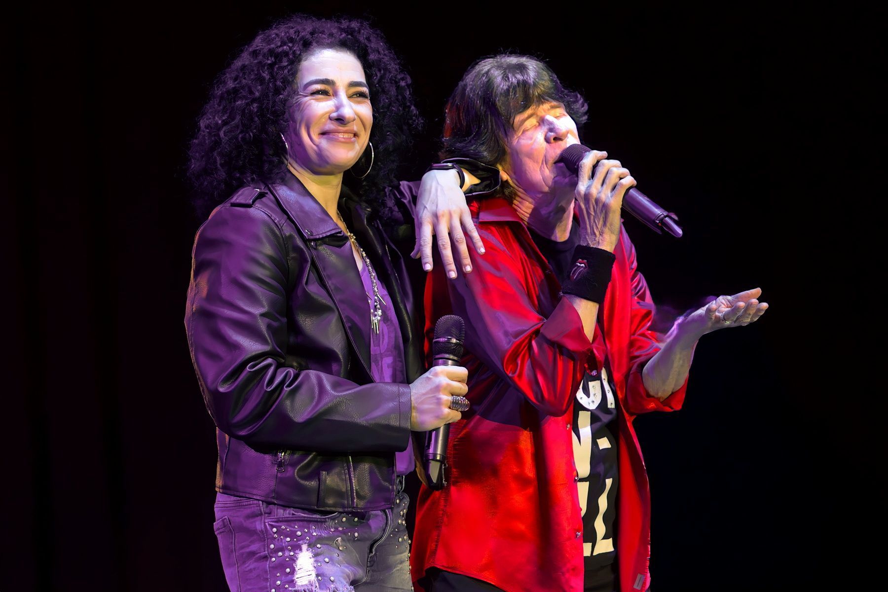 Two performers on stage, one singing into a microphone while the other smiles beside them.