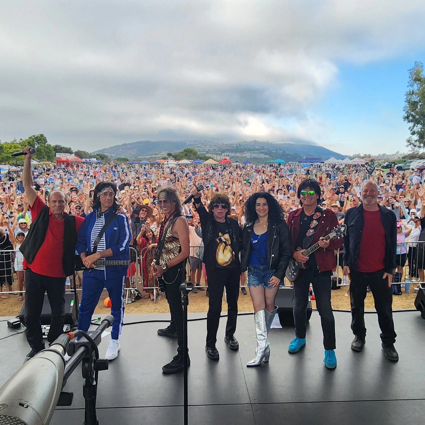 Six performers on a stage facing a huge outdoor crowd under cloudy skies and mountains.