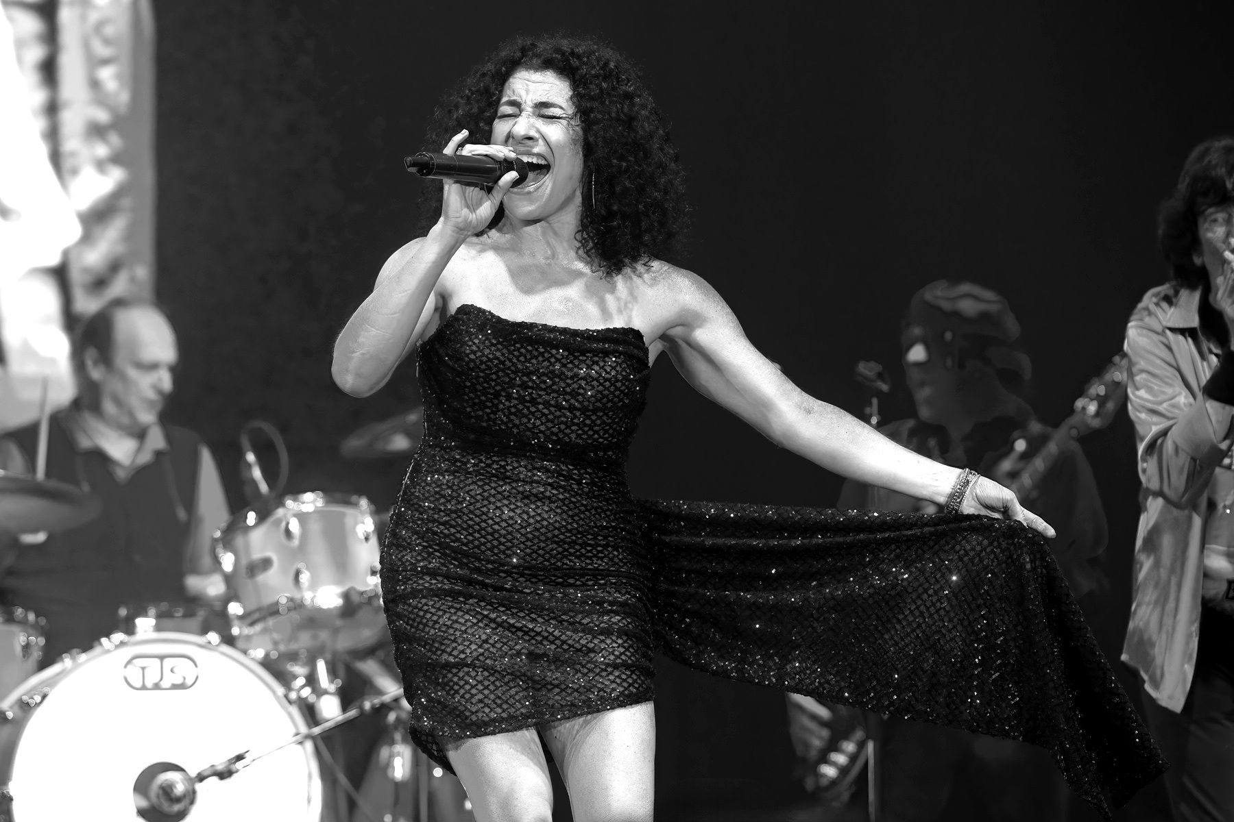 Singer performing on stage in a black dress, singing into a microphone with a live band behind her.